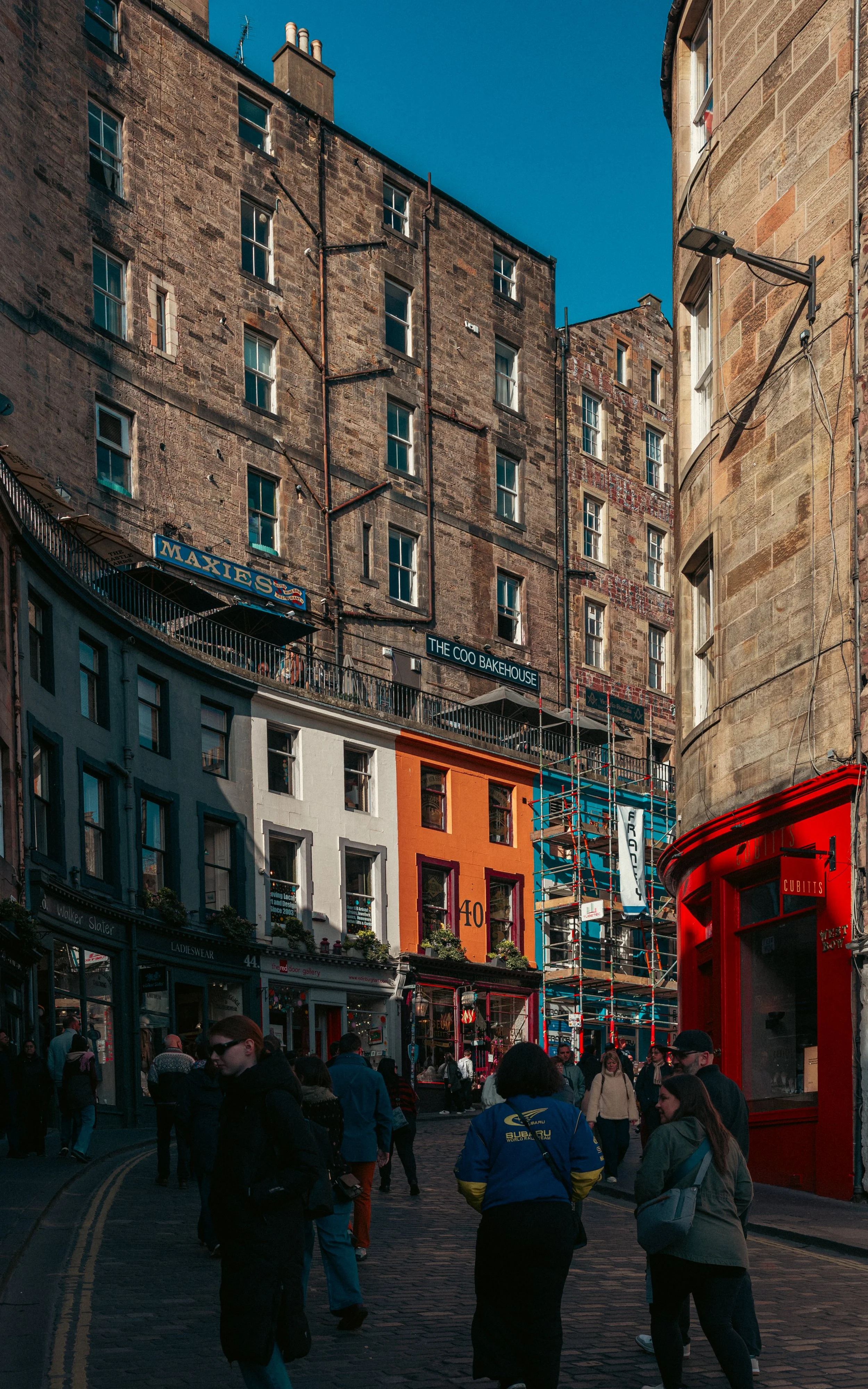 A busy city street with people walking near colorful storefronts, surrounded by tall brick buildings under a clear blue sky.