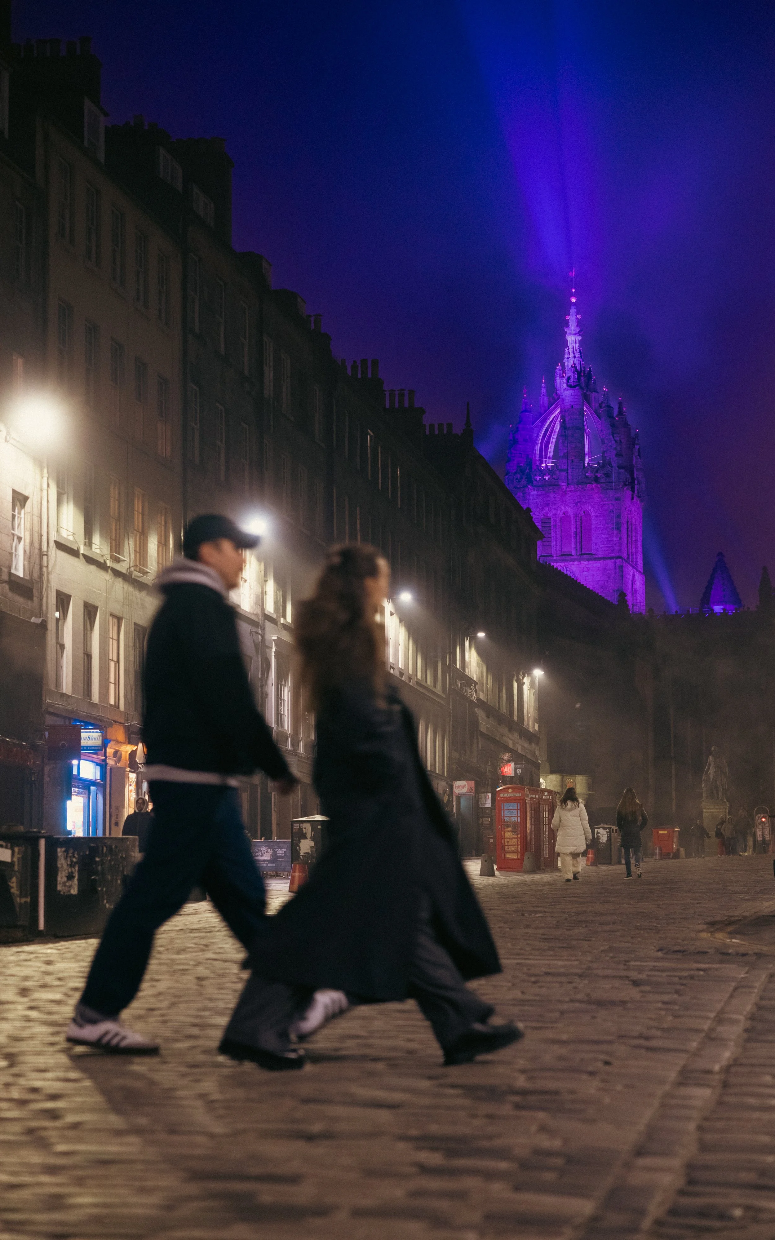 People walking on a cobblestone street at night, with historic buildings and a illuminated tower in the background.