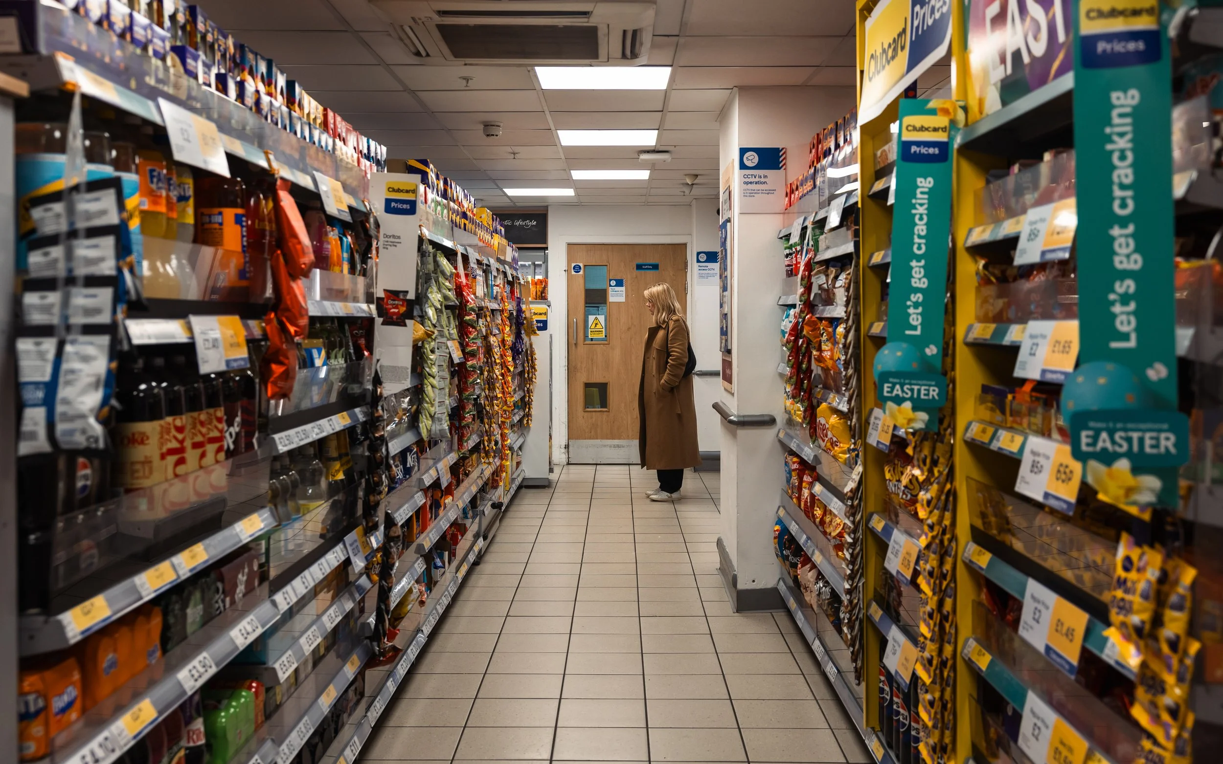 A woman in a brown coat and black pants stands in an aisle of a grocery store, looking at shelves filled with snacks and candies on either side. The store has a tiled floor, fluorescent lighting, and a wooden door at the end of the aisle.
