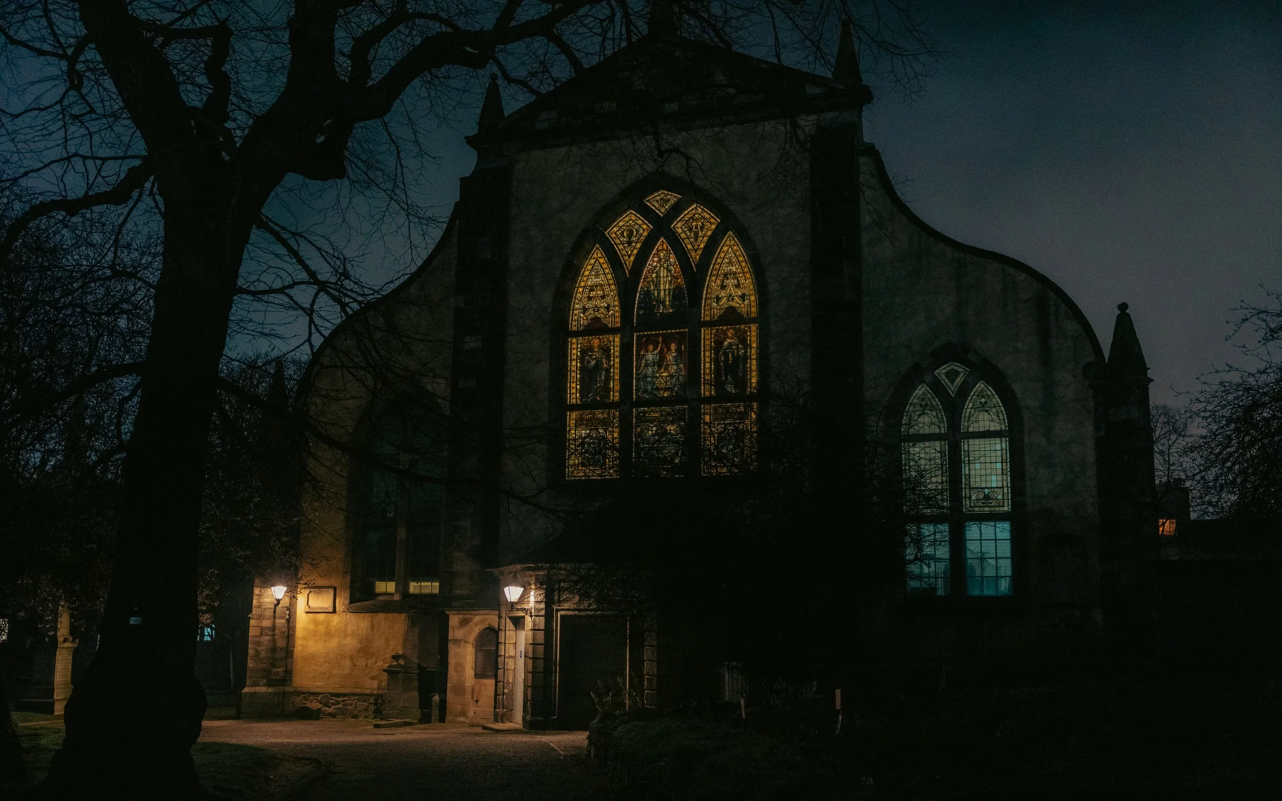 Nighttime view of a historic church with stained glass windows illuminated from inside, surrounded by leafless trees and lit street lamps.
