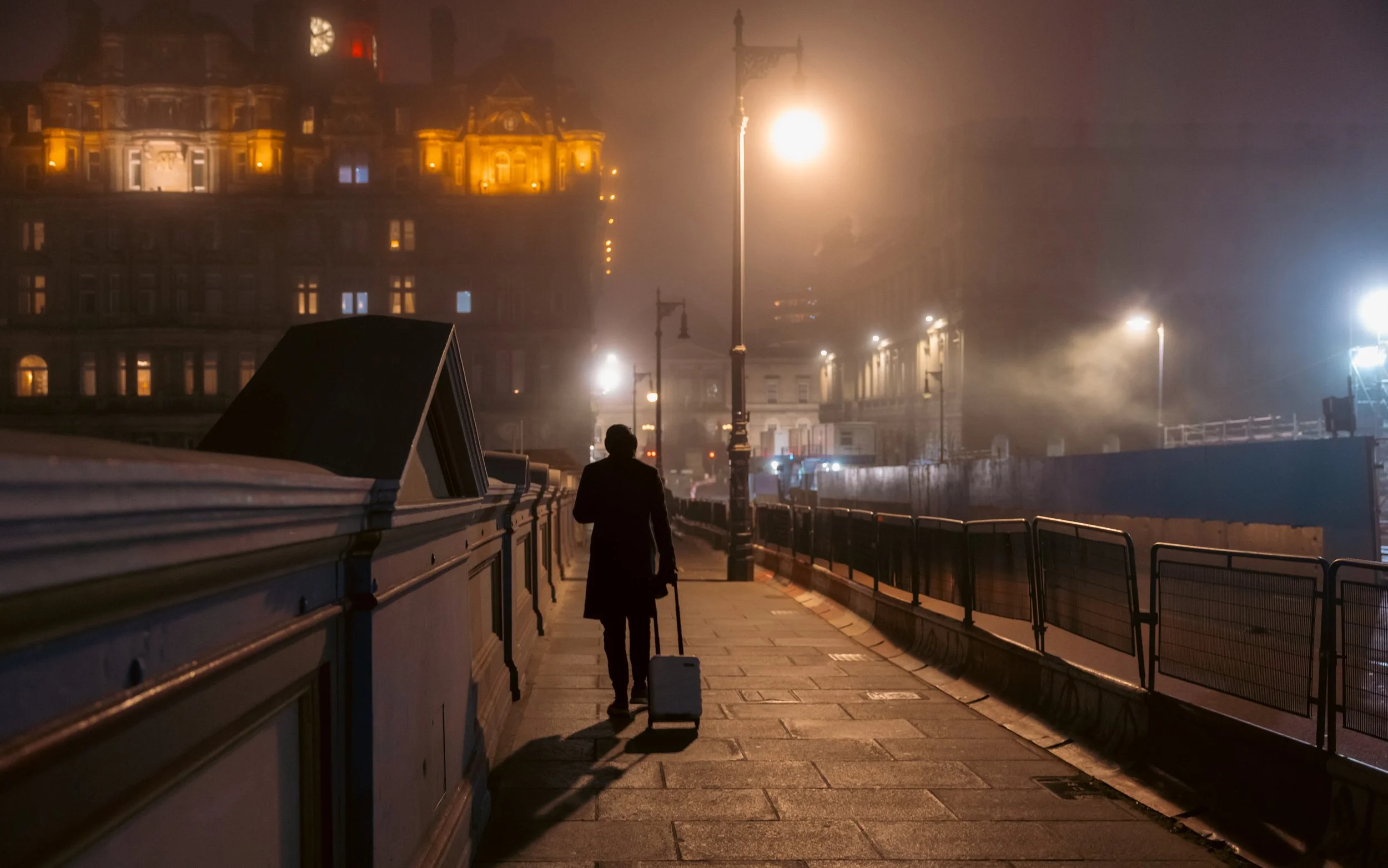 A person walking on a city sidewalk at night, pulling a suitcase, with fog and illuminated buildings in the background.
