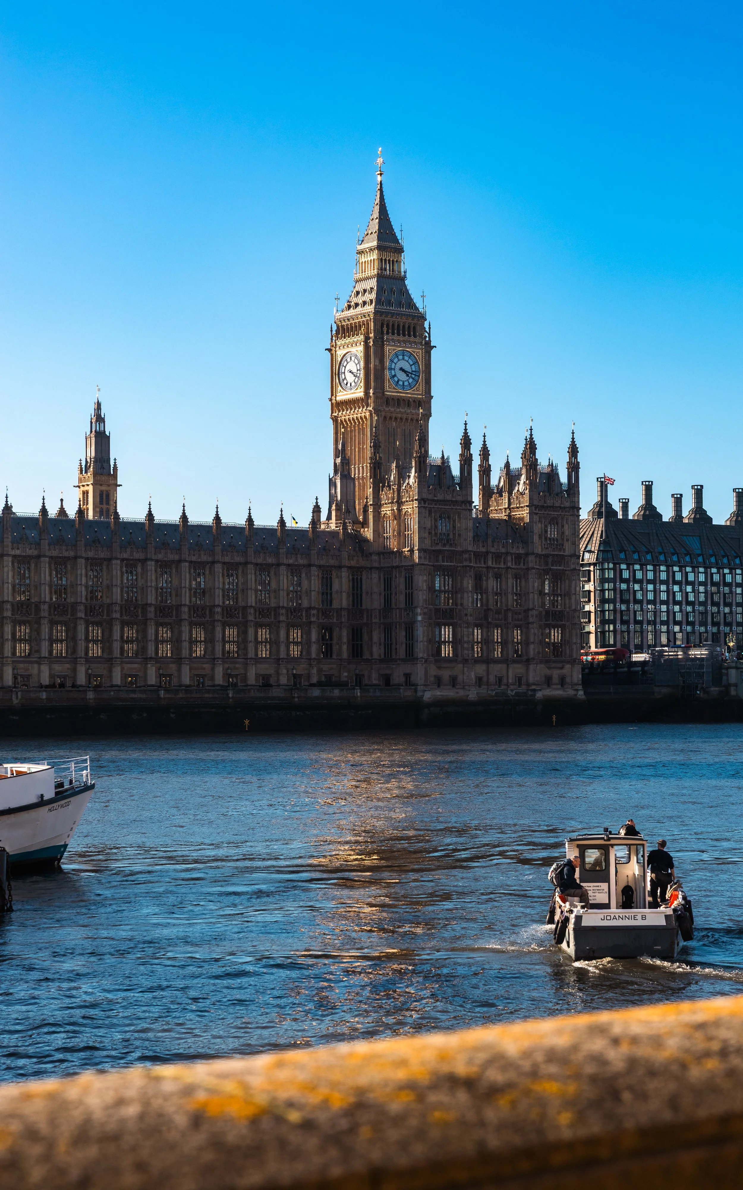 A view of the Big Ben clock tower and the Palace of Westminster in London, with a boat on the river Thames in the foreground and a clear blue sky above.
Taken in London