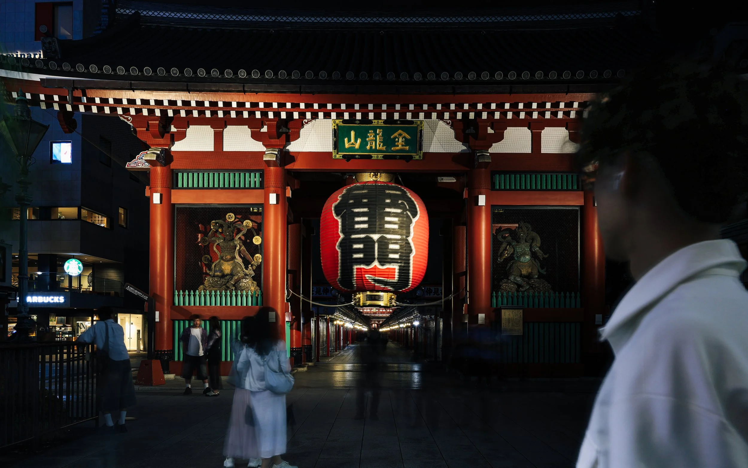 A night view of the Kaminarimon gate at Senso-ji Temple in Tokyo, Japan, featuring a large red lantern with black kanji characters, traditional statues, and illuminated surroundings with people walking near the entrance.