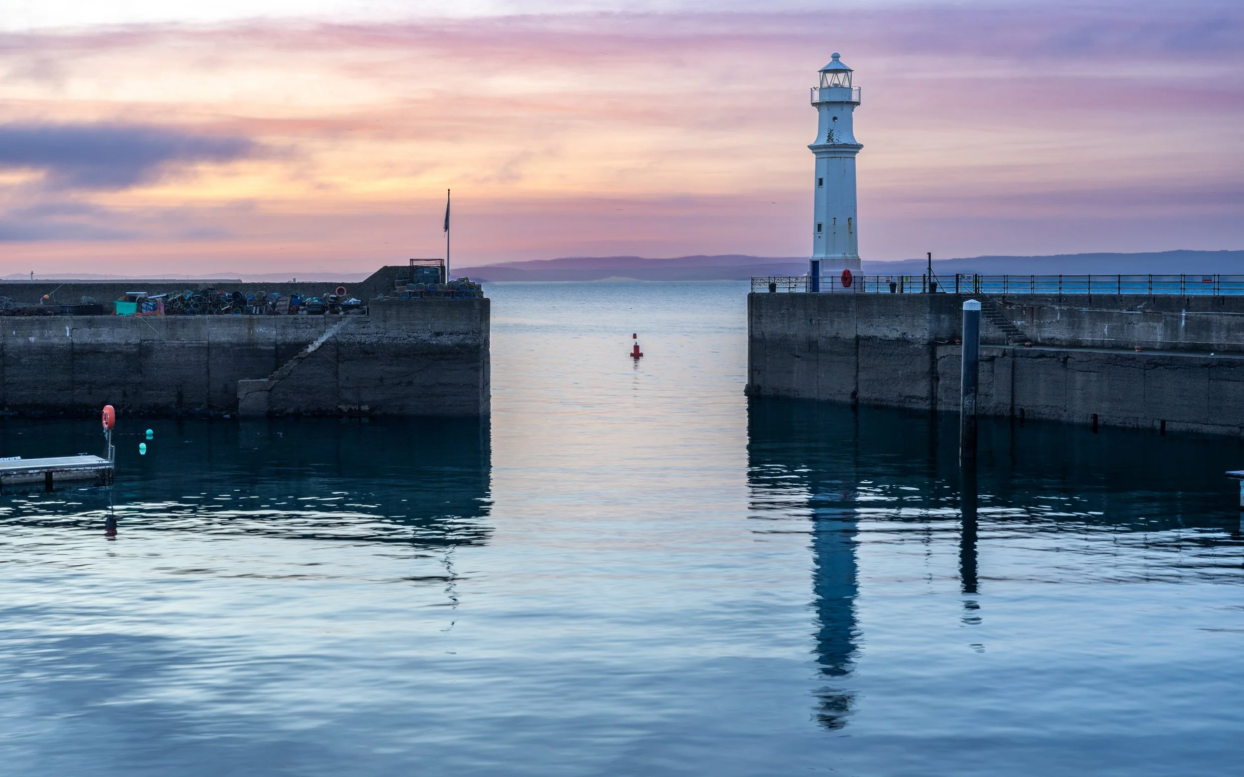 A lighthouse stands on a pier at sunset, with calm bluish water reflecting both the lighthouse and the pier against a violet-colored sky.