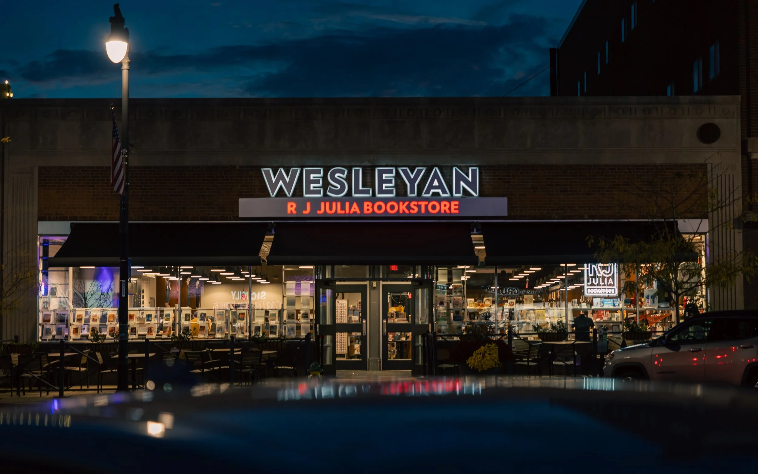 Night view of Wesleyan bookstore with illuminated sign, large glass storefront, and outdoor seating.