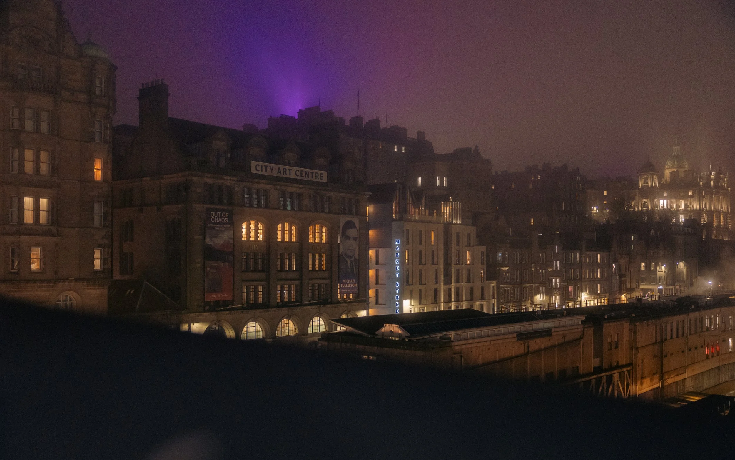 Night view of a city with illuminated buildings, including a sign that reads 'City Art Centre' and a vertical sign for 'Market Street', with purple lights in the sky.