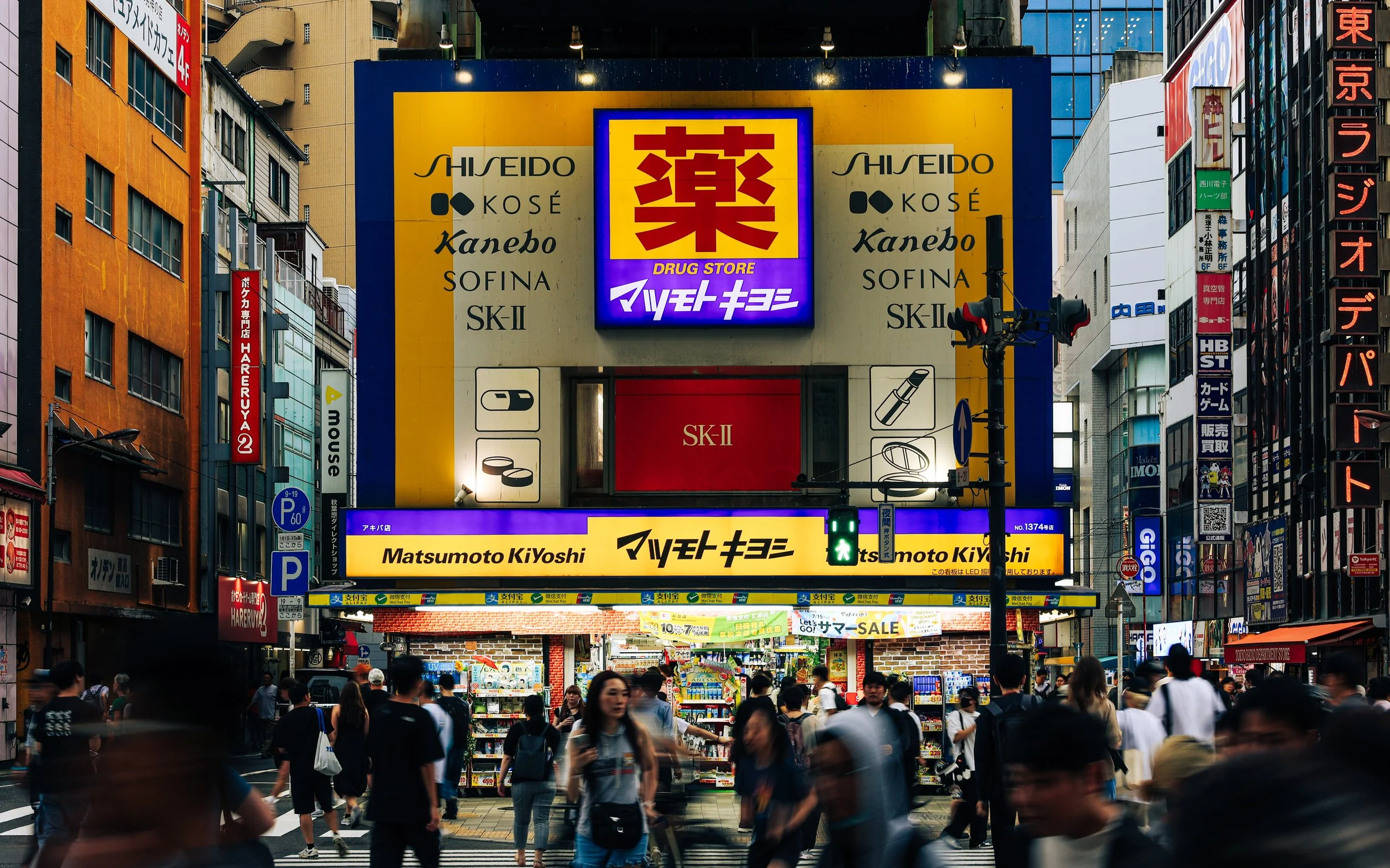 A busy city street scene in Japan with many pedestrians and bright advertisements, including a large yellow and blue sign for a drug store with Japanese characters, electronic billboards, and storefronts.