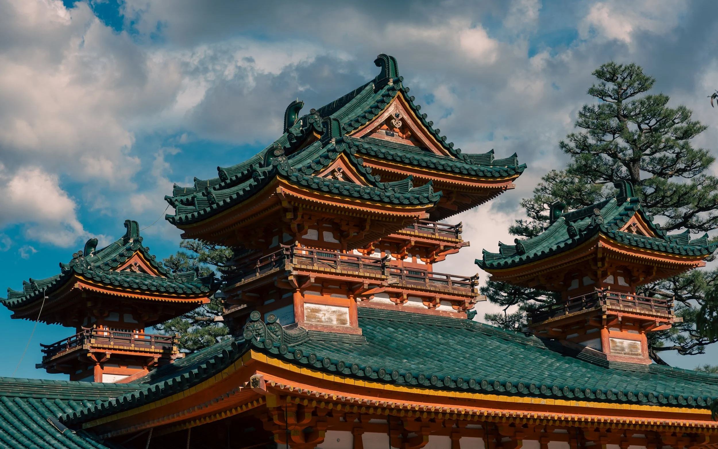 Traditional Japanese temple with tiered, green-tiled roofs and ornate woodwork, set against a partly cloudy sky and pine trees.