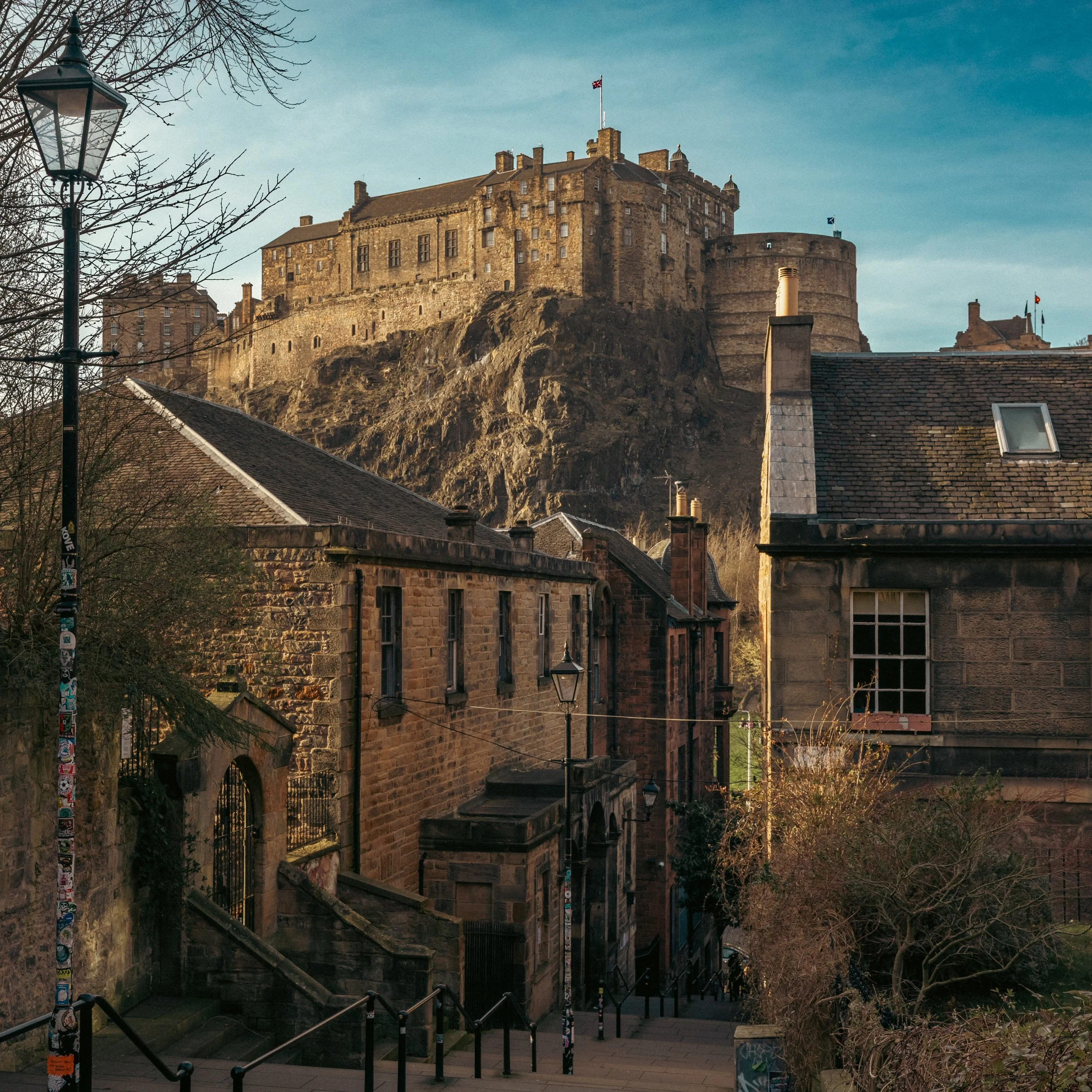View of Edinburgh Castle on a hill overlooking old buildings and a street with lampposts and stairs.