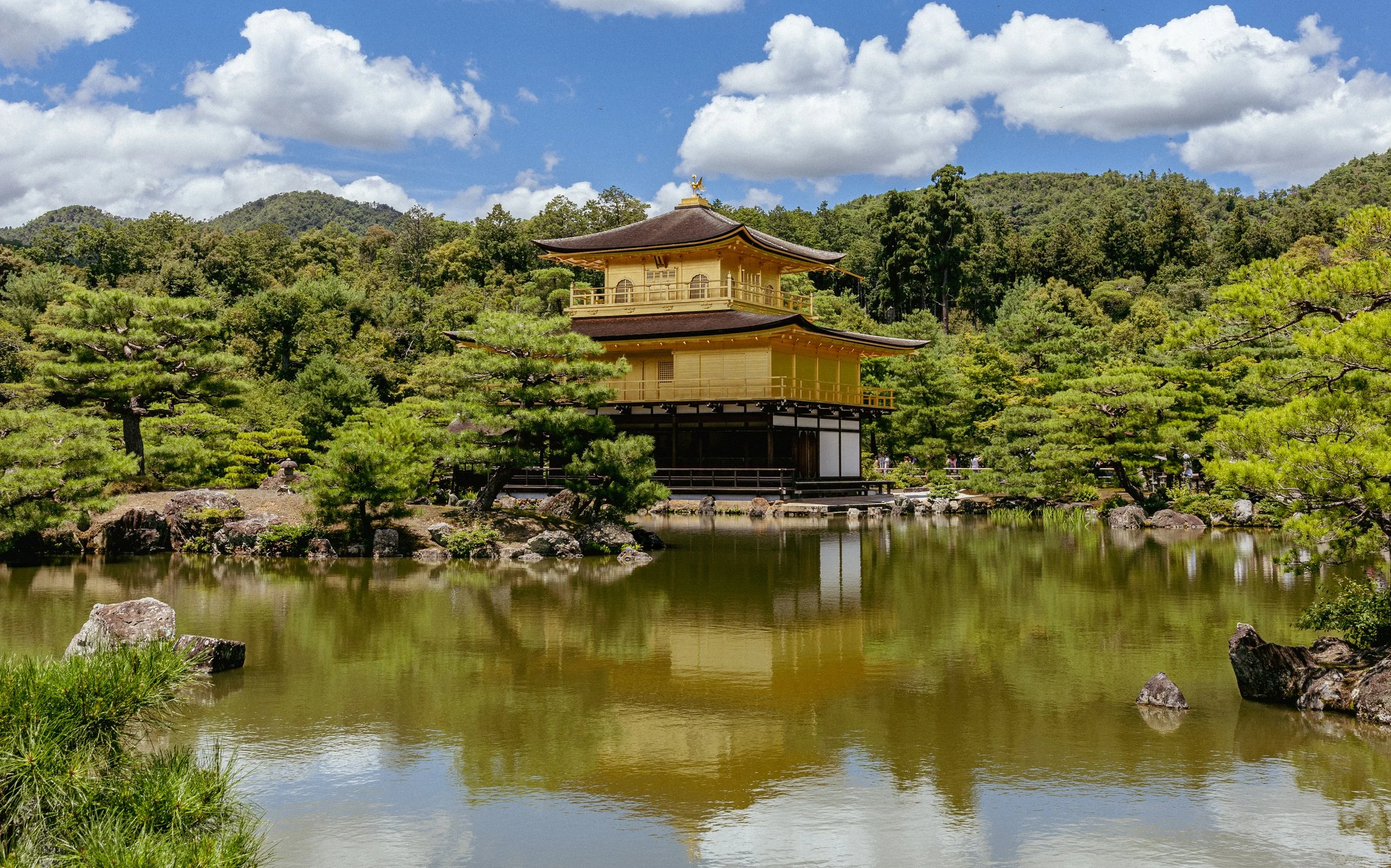 Kinkaku-ji, or the Golden Pavilion, reflecting in a pond surrounded by trees and green hills.