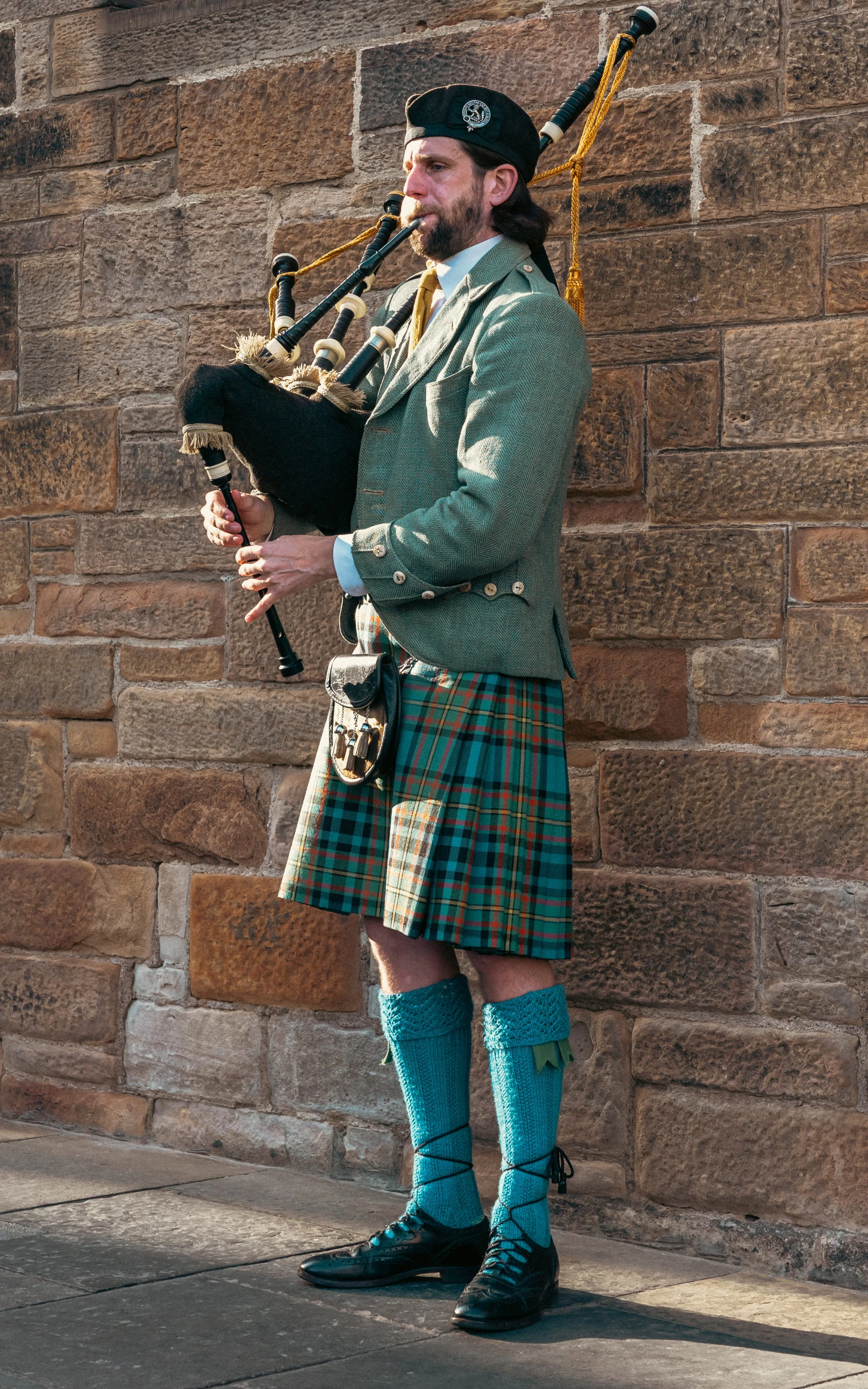 A man dressed in traditional Scottish attire playing bagpipes against a brick wall.