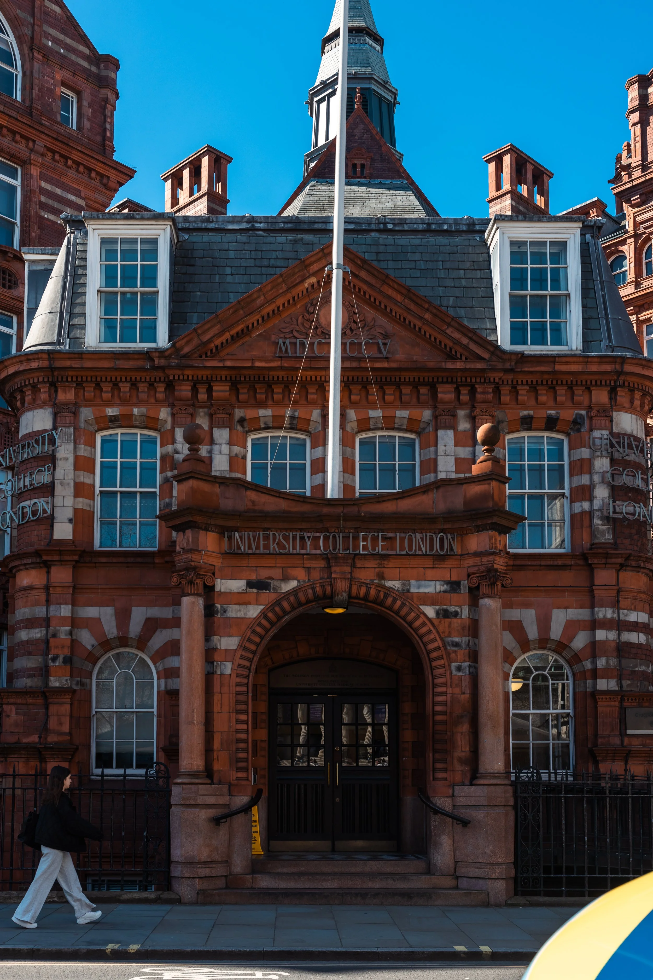 Red brick building with arched windows and entrance, sign reading 'University College London,' and a flagpole in the front. A person with long hair and a backpack walking on the sidewalk.