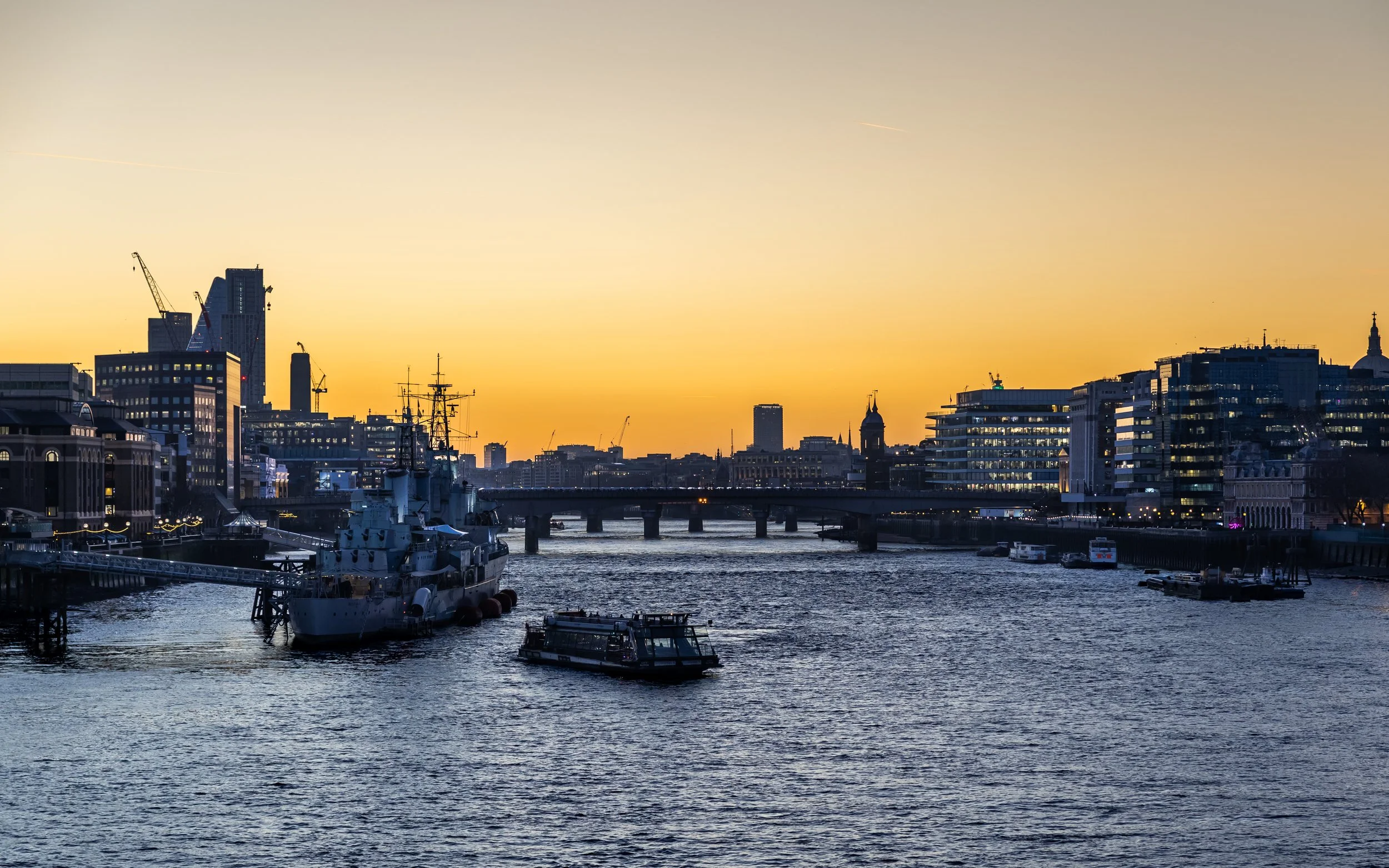Sunset over a city river with boats, modern buildings, and a bridge.
Taken in London