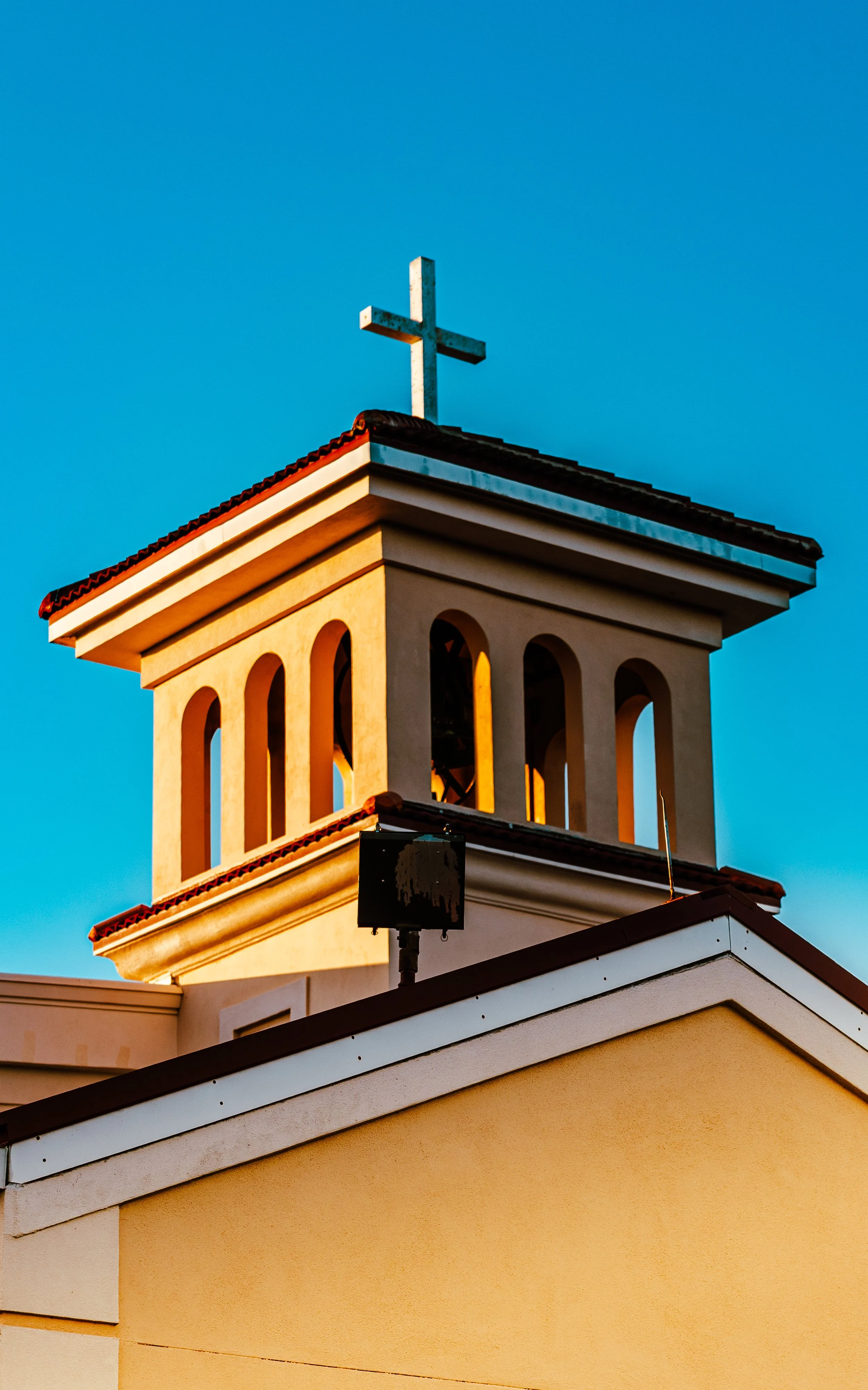 Church steeple with a cross on top, illuminated by warm sunlight, set against a clear blue sky.