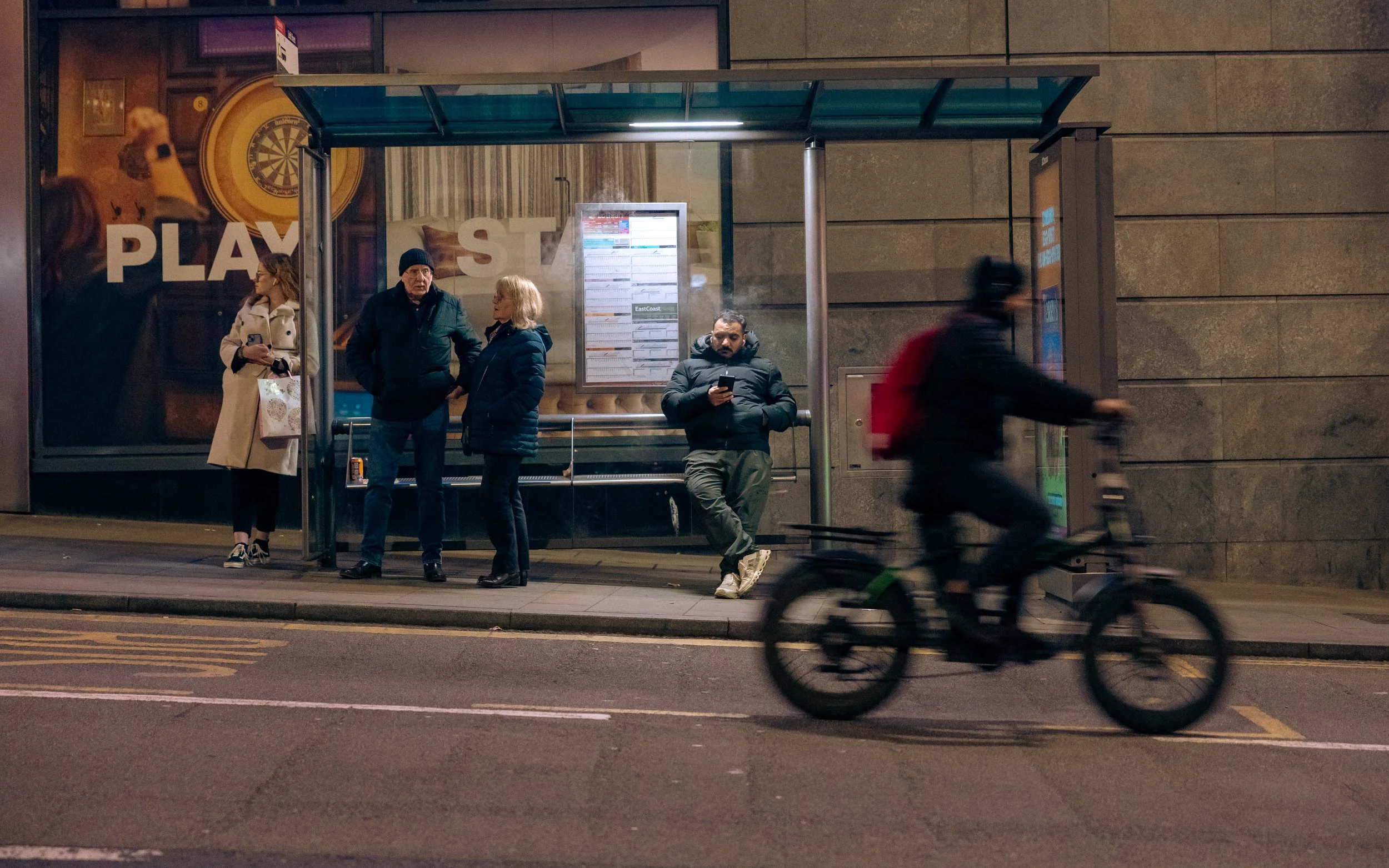 A city bus stop with four people waiting, three standing and one sitting, and a cyclist passing by in the foreground at night.