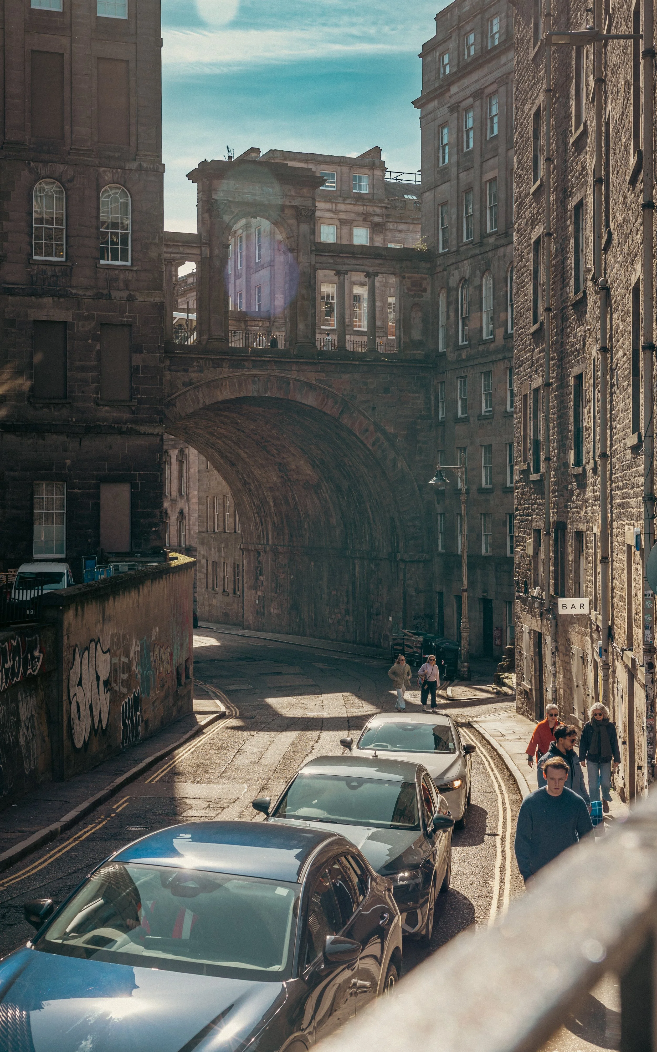 Street scene with cars parked along a narrow road, pedestrians walking, historic stone buildings, an arch bridge, and graffiti on a wall, under a partly cloudy sky.