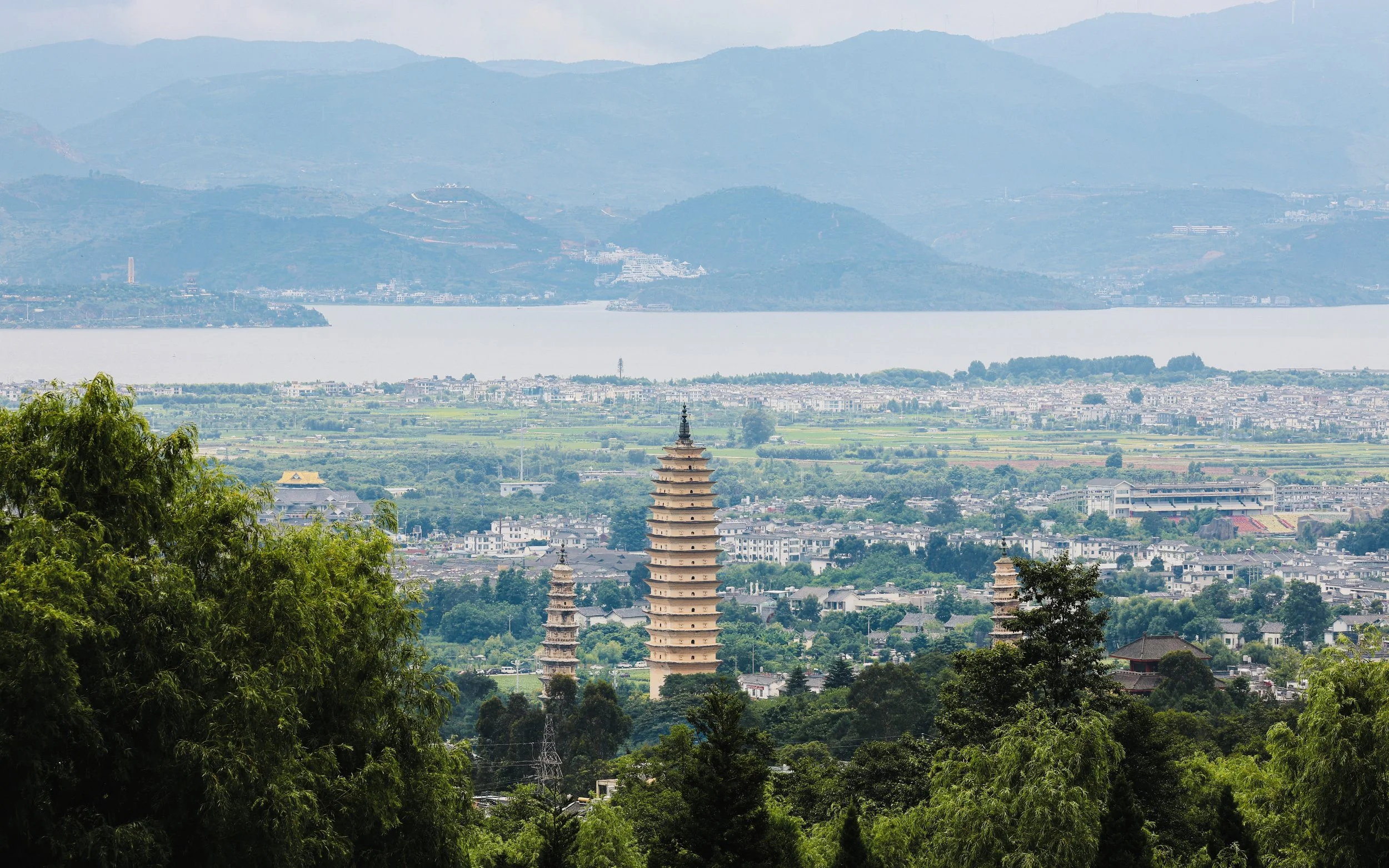 Landscape view of lush green trees in the foreground, with three pagodas and an urban area beyond, and mountains and a lake in the background.