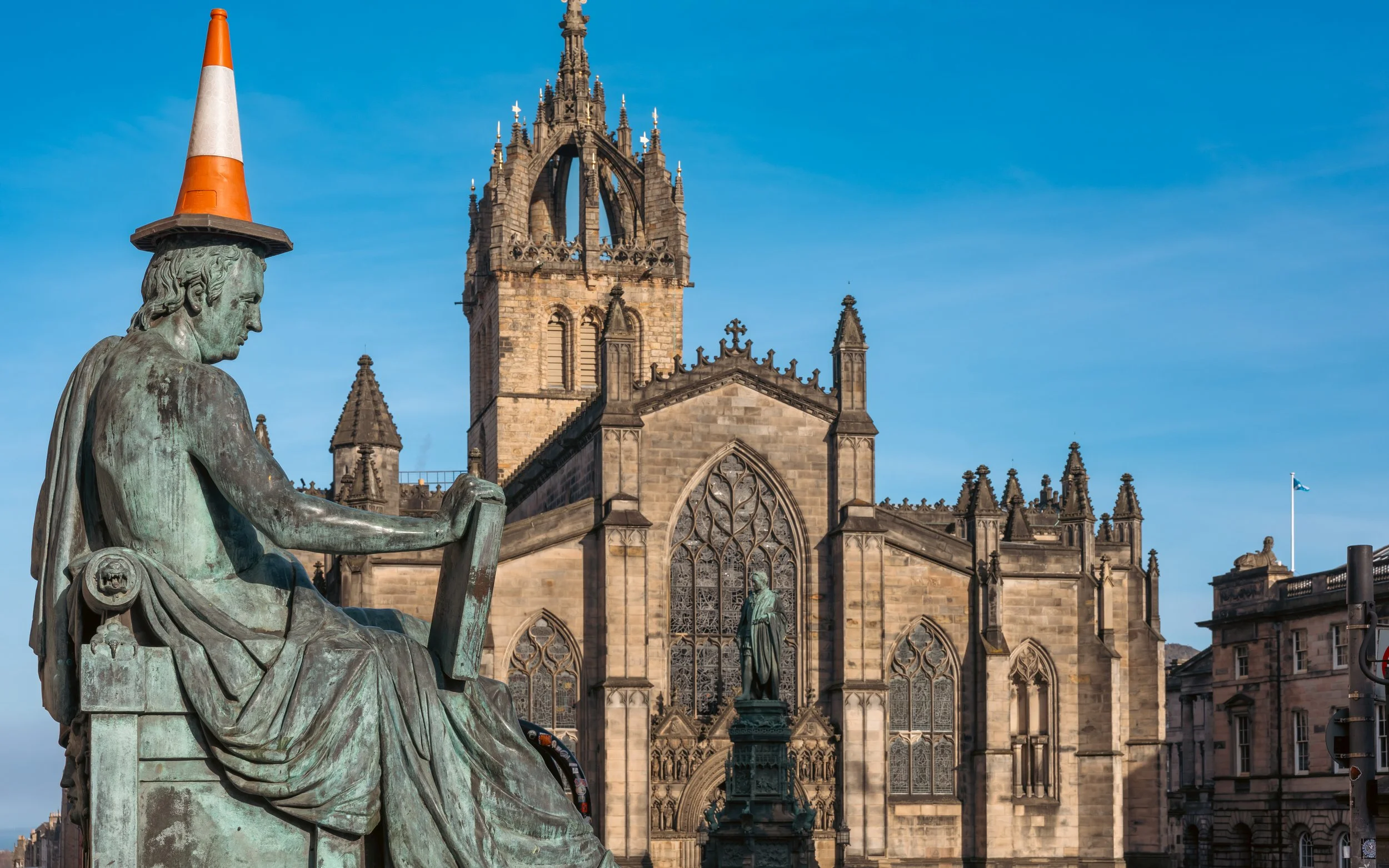 Bronze statue of a seated figure holding a scroll, with an orange and white traffic cone on its head, in front of a Gothic-style cathedral with a tall clock tower and stained glass windows, under a blue sky.