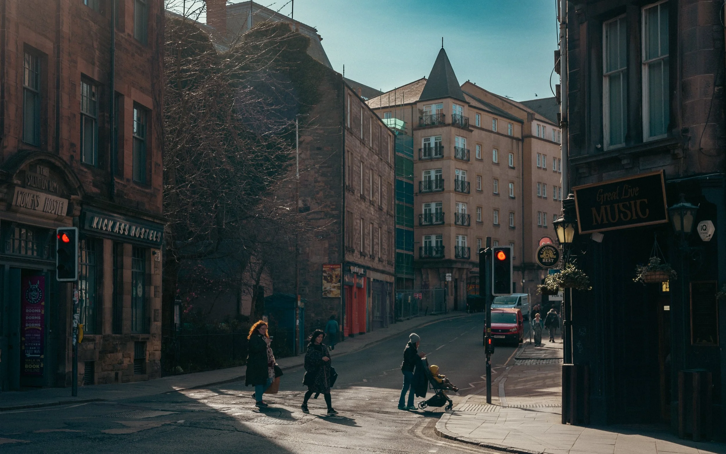 People walking cross a city street with traffic signals, buildings, and shops in the background, in a sunny day.
