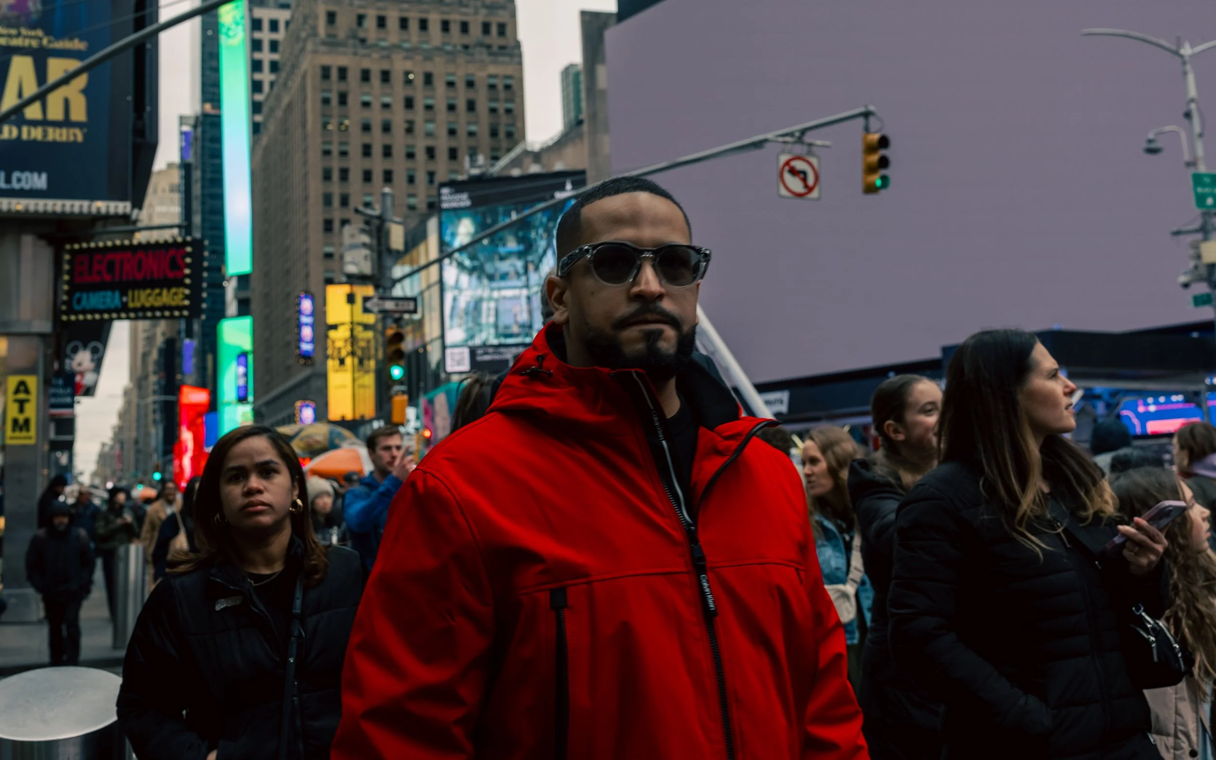 Man in red jacket and sunglasses standing among a crowd in Times Square, New York City, with bright lights and digital billboards in the background.