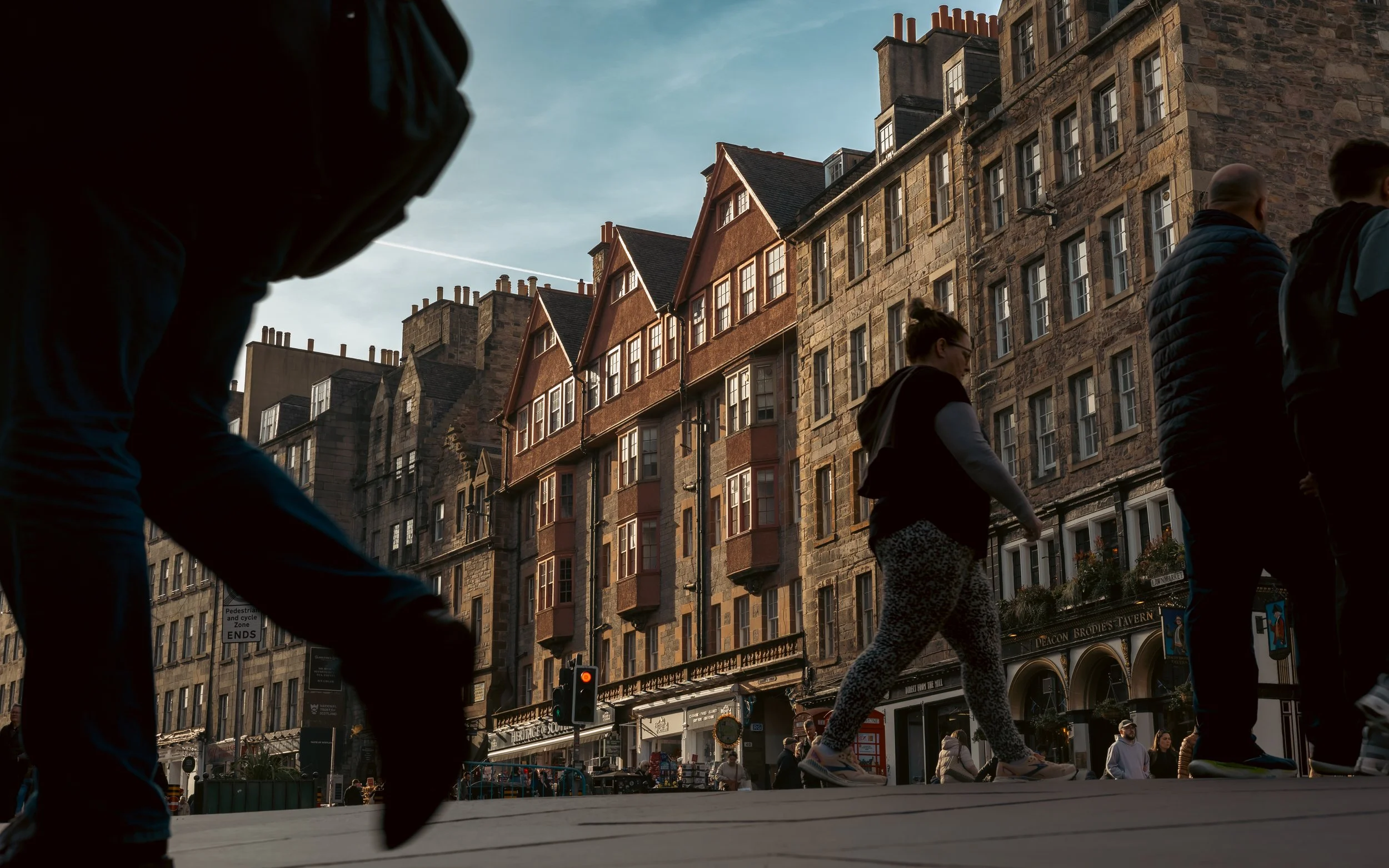 People walking on a city street in front of historic stone and brick buildings with multiple windows and protruding bay windows, during daytime with a clear sky.