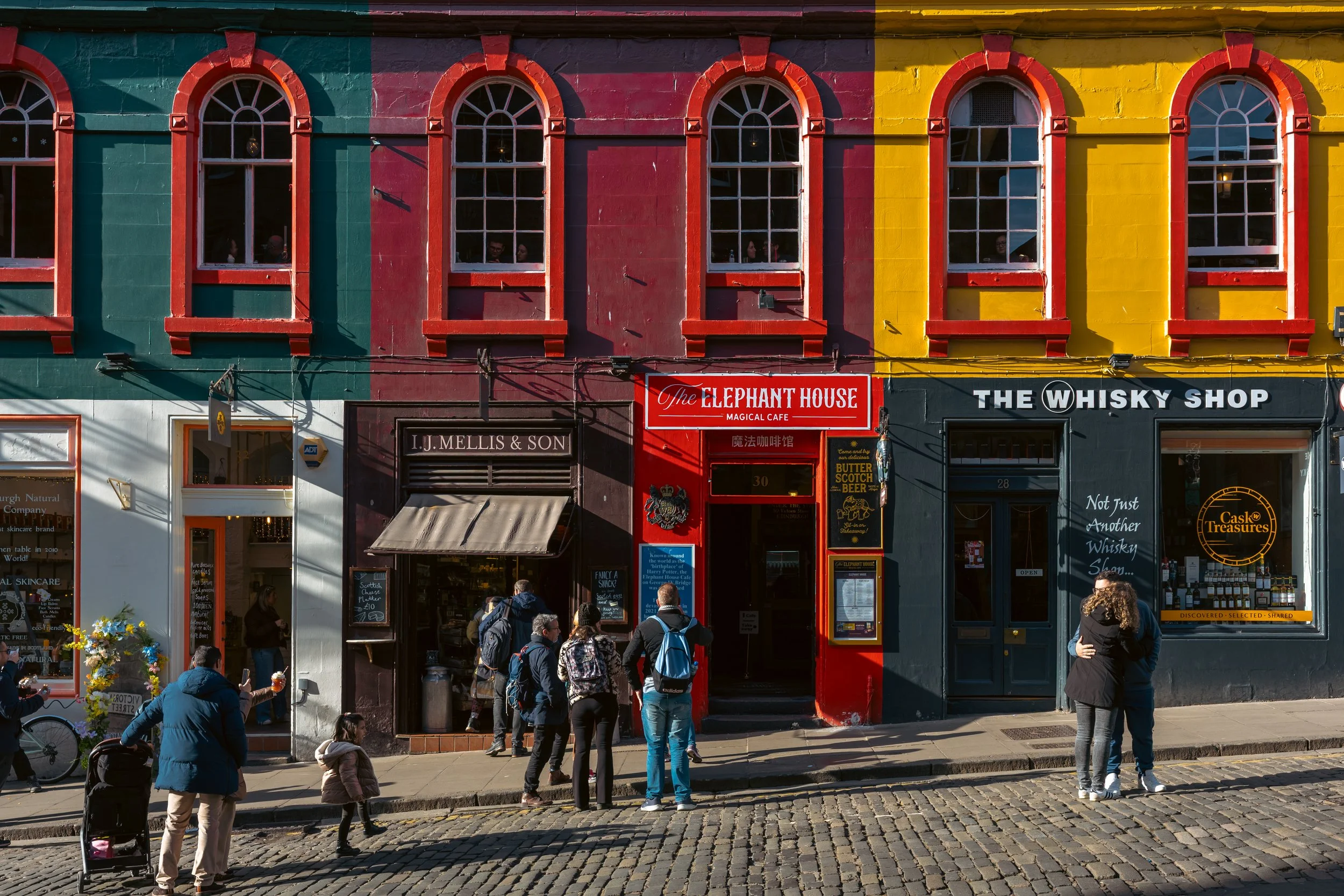 Colorful buildings with red, green, yellow, and black facades, with people walking and standing on the cobblestone street outside shops and cafes.