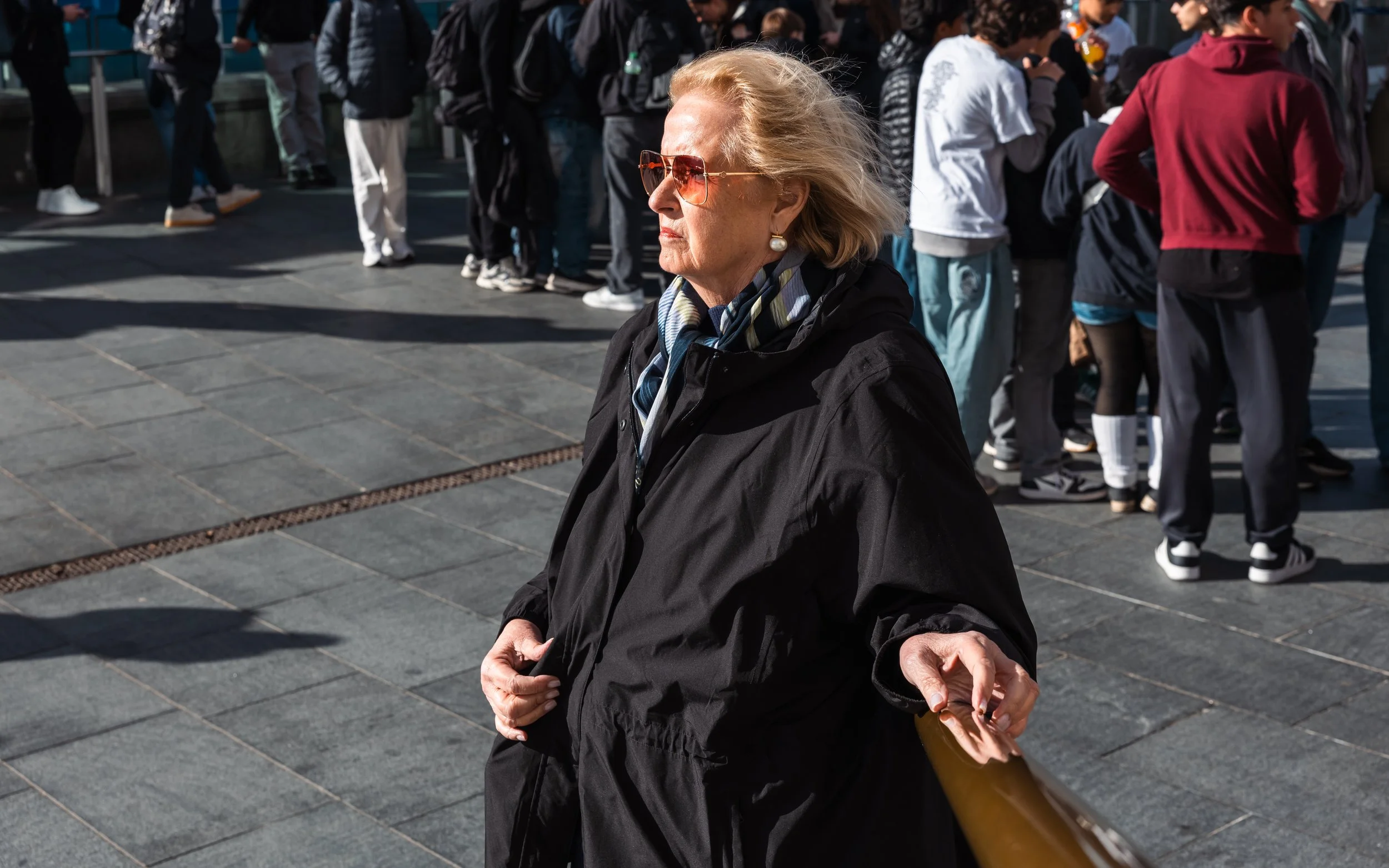 An older woman with blonde hair, wearing sunglasses, a black coat, and a scarf, standing on a busy outdoor urban area with a crowd of people in the background.