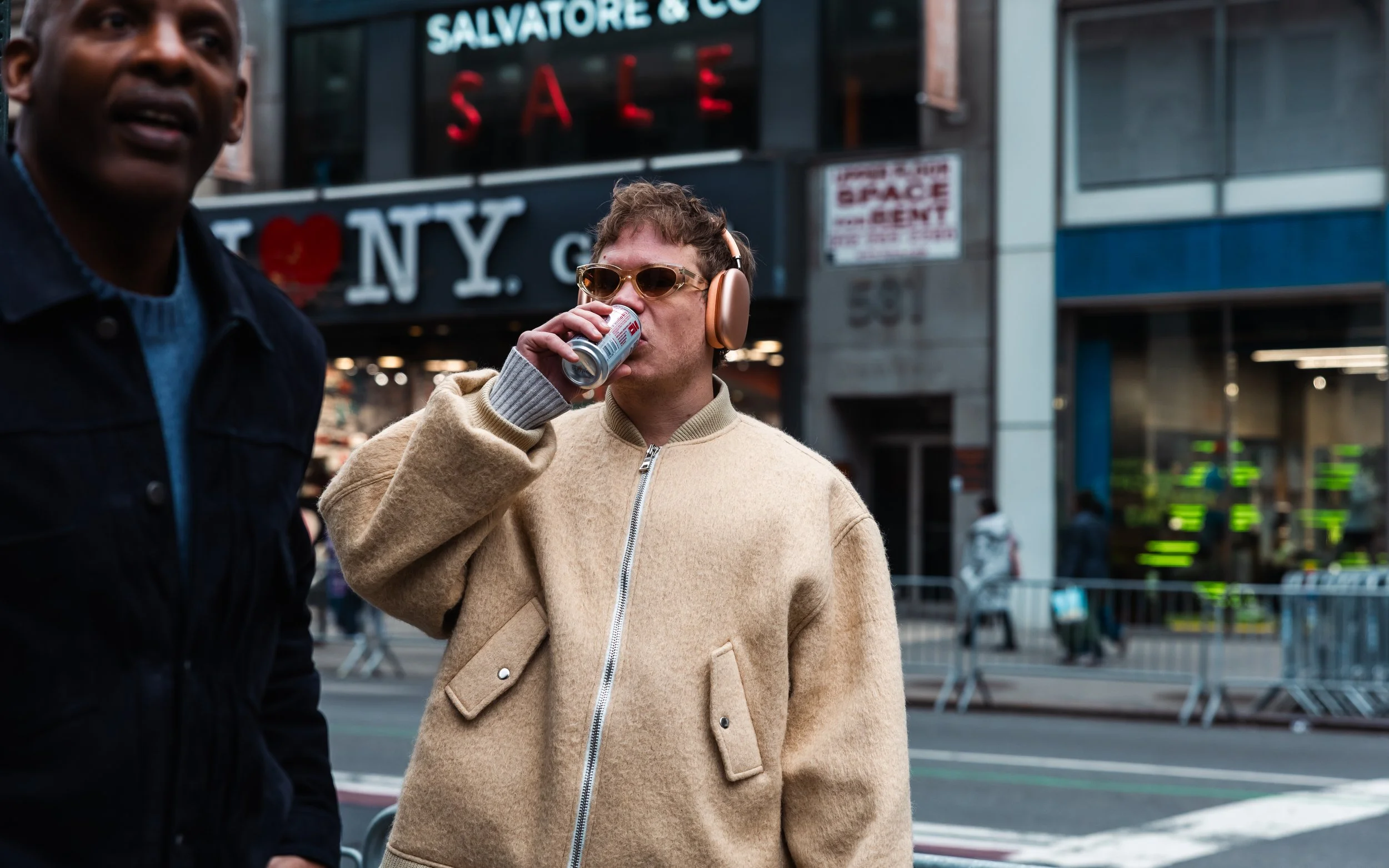 A person with short hair wearing sunglasses, beige jacket, and headphones, drinking from a can on a city street, with NY signage and a store in the background.