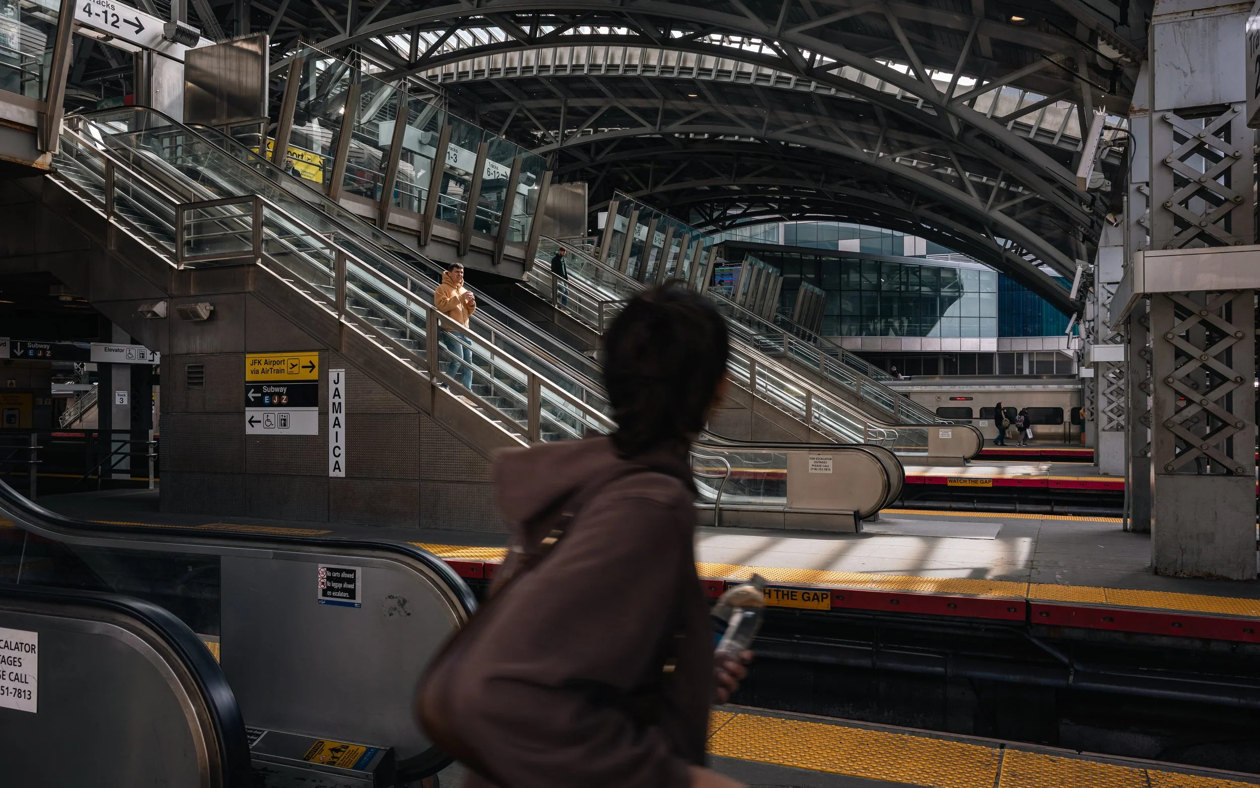 A subway station with escalators and signs. A person in the foreground is holding a phone, facing away from the camera. Another person is on the escalator, and a few more are walking across the platform. The station is modern with glass and steel arc