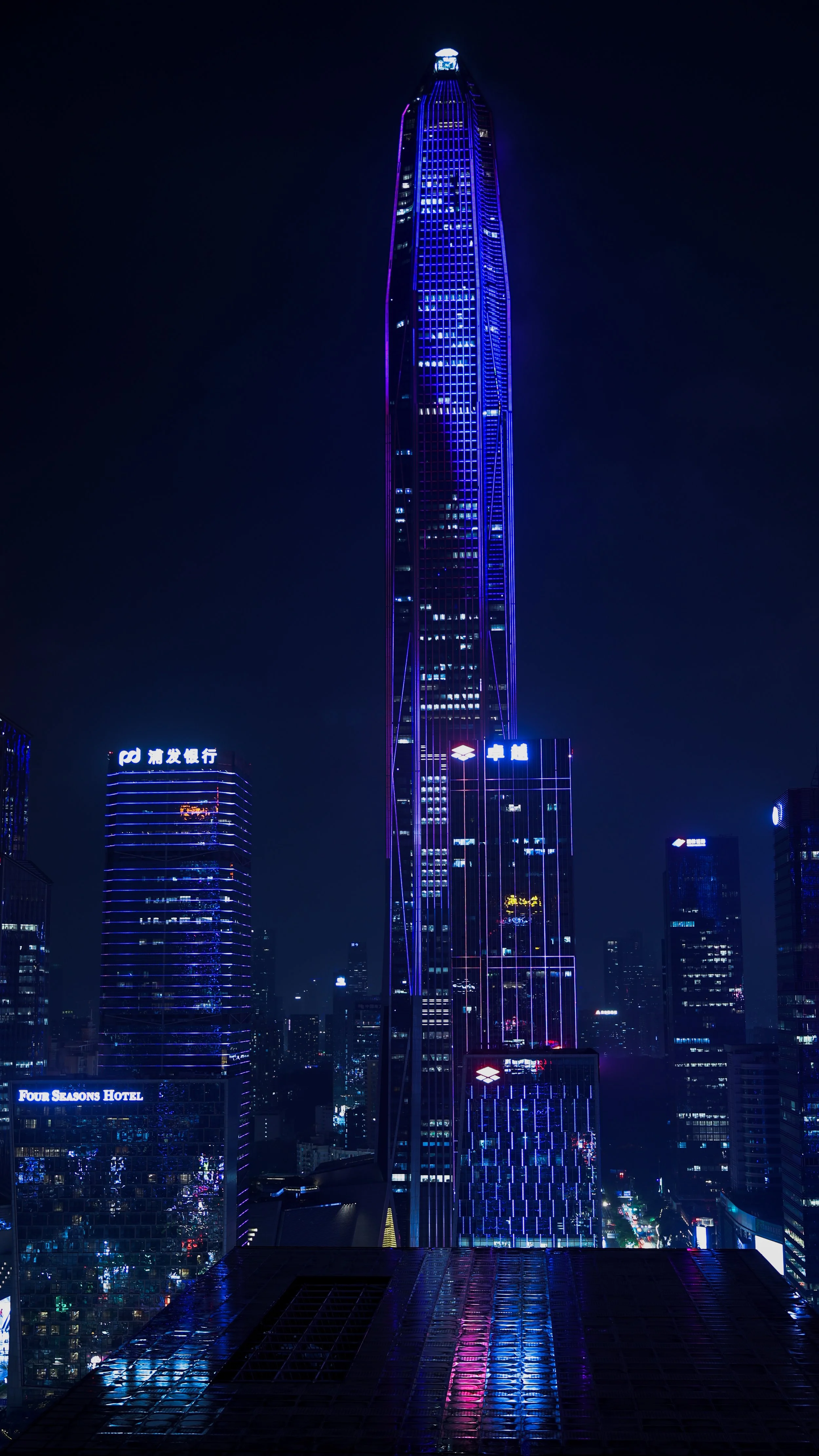 Nighttime view from Shenzhen Futian district with illuminated skyscrapers, including a tall, narrow building with blue lighting and a rounded top.