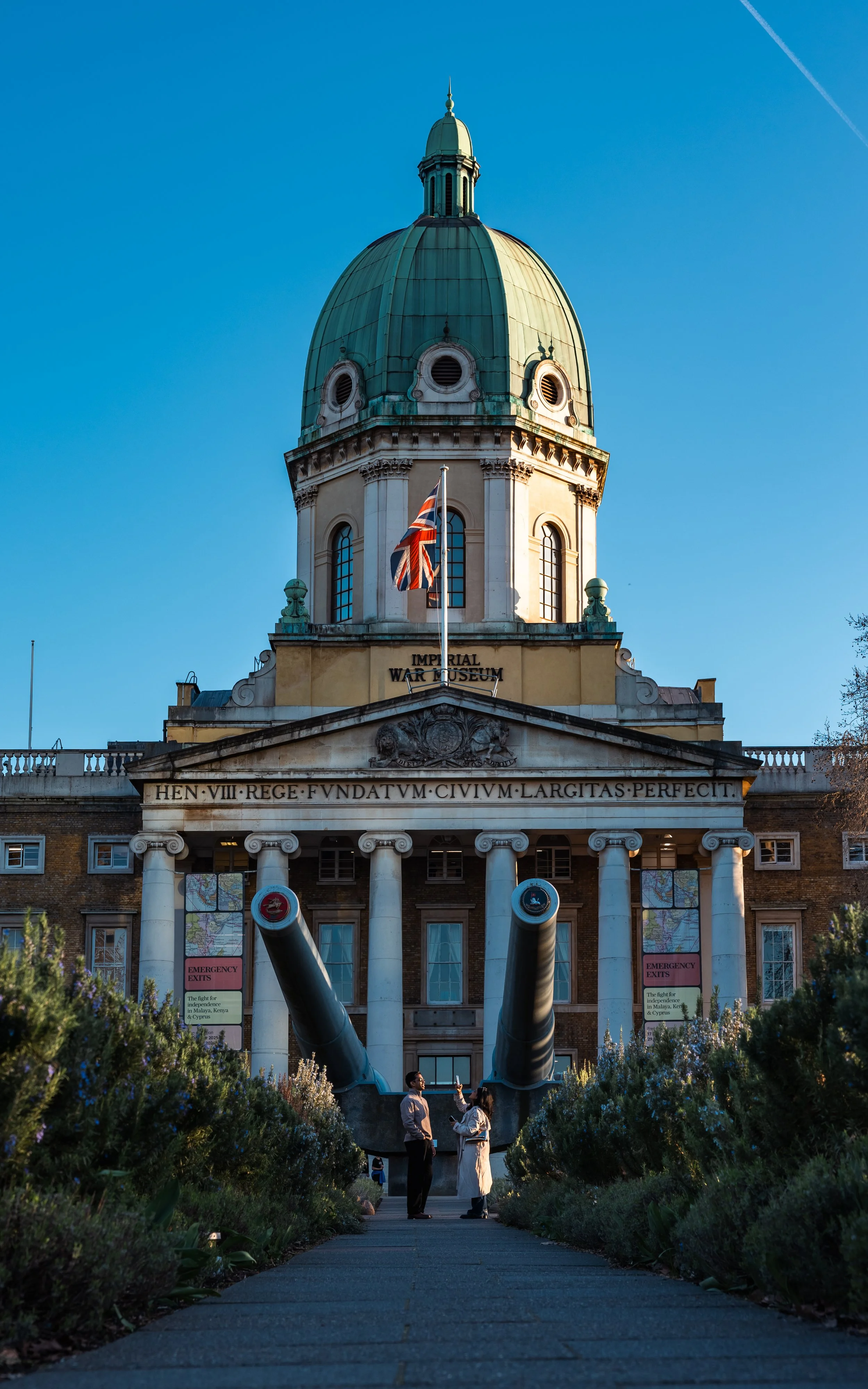 A man and woman standing in front of the artillery piece, with the Imperial War Museum in the back. 
Taken in London