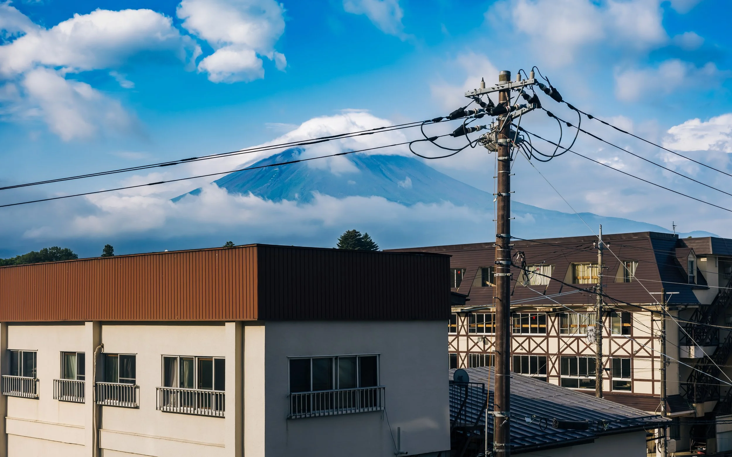 Cityscape with utility poles and power lines in foreground, buildings with balconies, and Mount Fuji covered in clouds in the background.