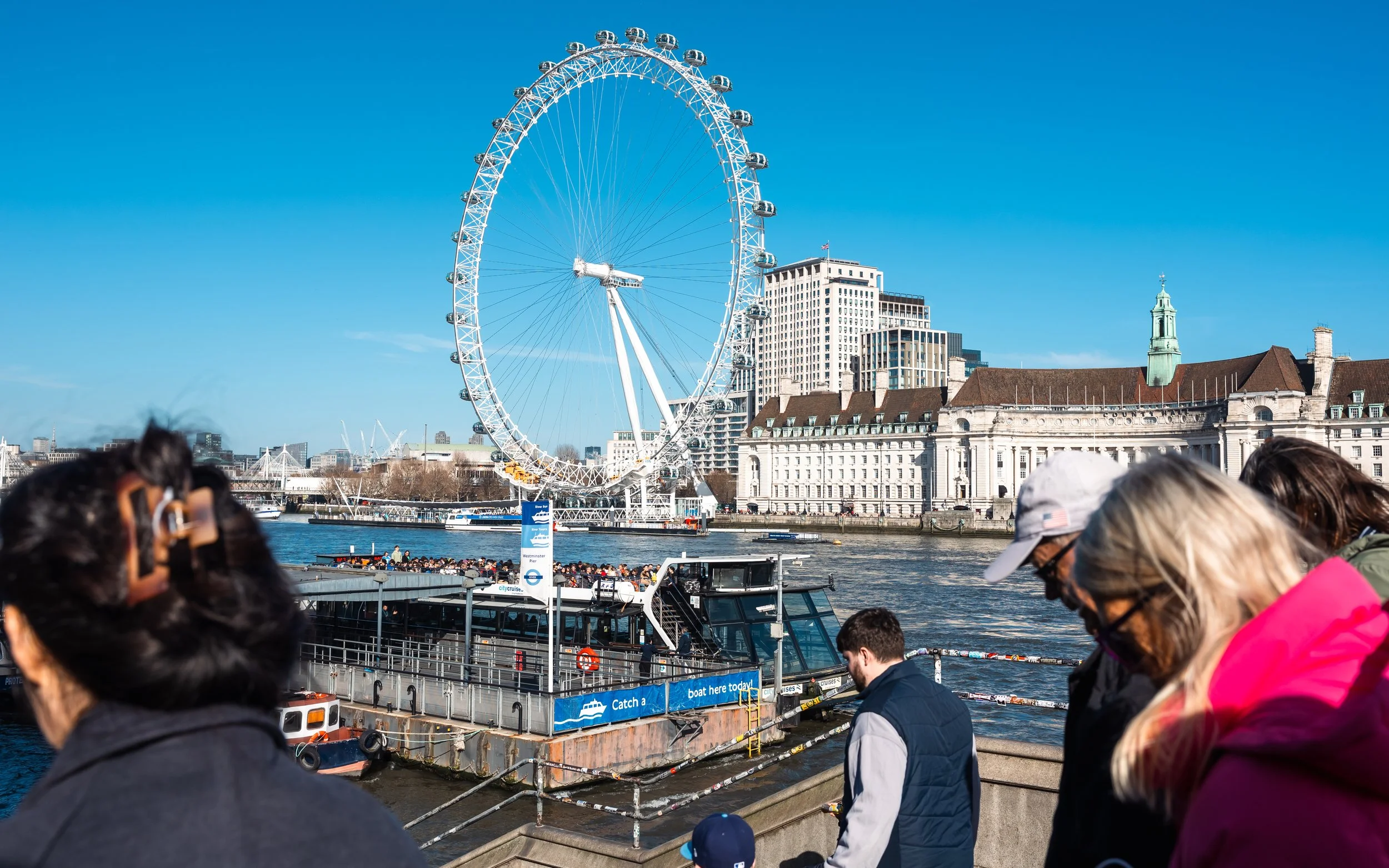 Walking pedestrians in the foreground, with the London Eye, calmly stand along the riverbank under a clear blue sky.
Taken in London