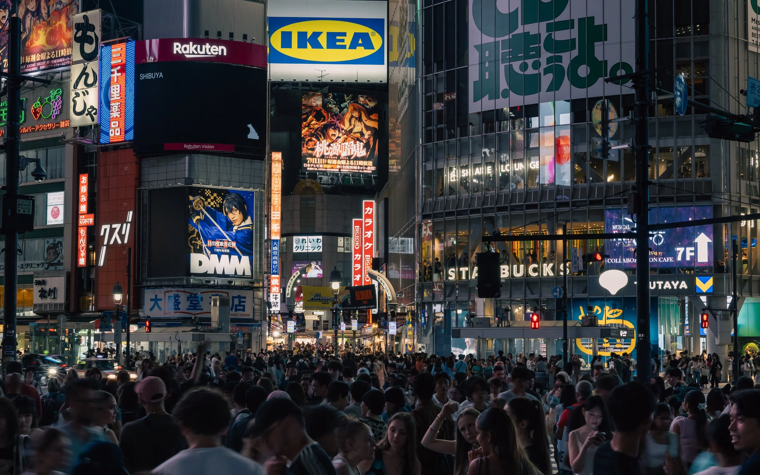 A busy city intersection at night filled with crowds of people and illuminated billboards in Japanese and English, including an IKEA sign, Starbucks, and various advertisements.