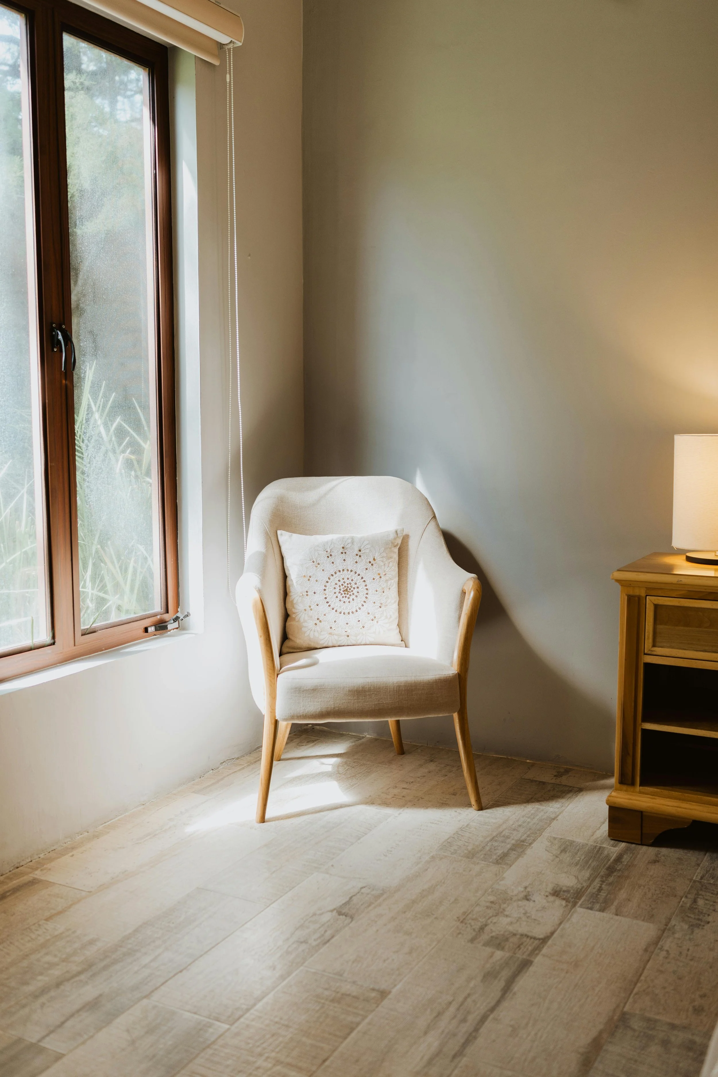 A cozy corner of a room with a beige armchair with wooden legs and armrests, a decorative pillow, a wooden side table with a lamp, and a window with natural light coming through.