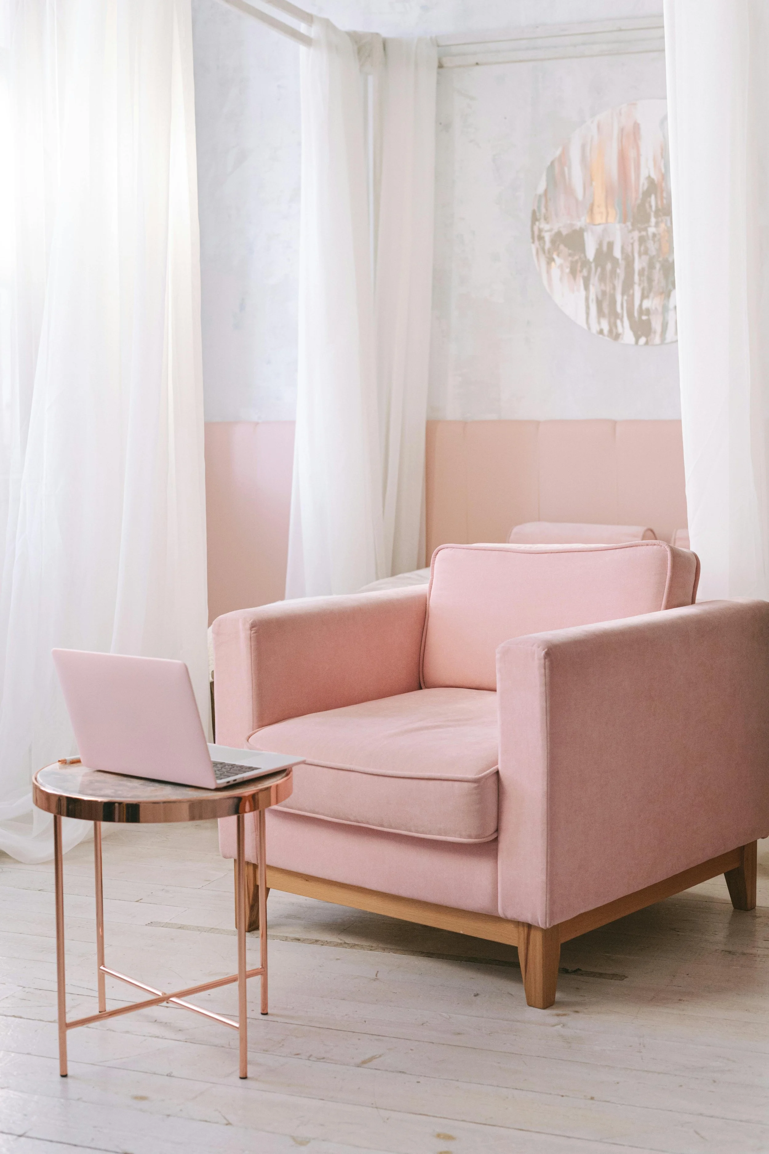 A pink armchair with a cushion, a small round side table with a laptop on it, set in a light, airy room with white curtains and abstract art on the wall.