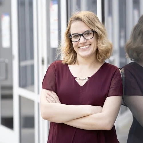 Smiling woman with blonde hair, glasses, and a maroon dress standing outside near glass doors.