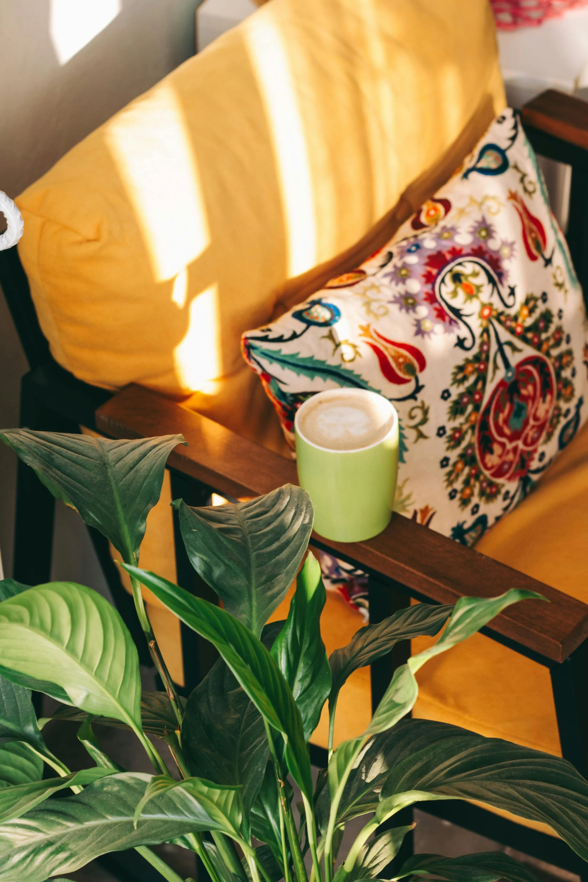 A cozy indoor scene with a yellow sofa, a colorful embroidered pillow, a green ceramic mug of coffee, and a large green leafy plant.