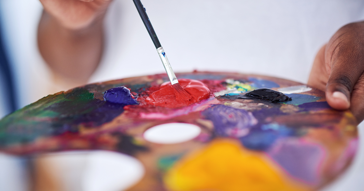 Close-up of an artist's hand holding a palette with various colors of paint, and a small brush dipped in red paint.