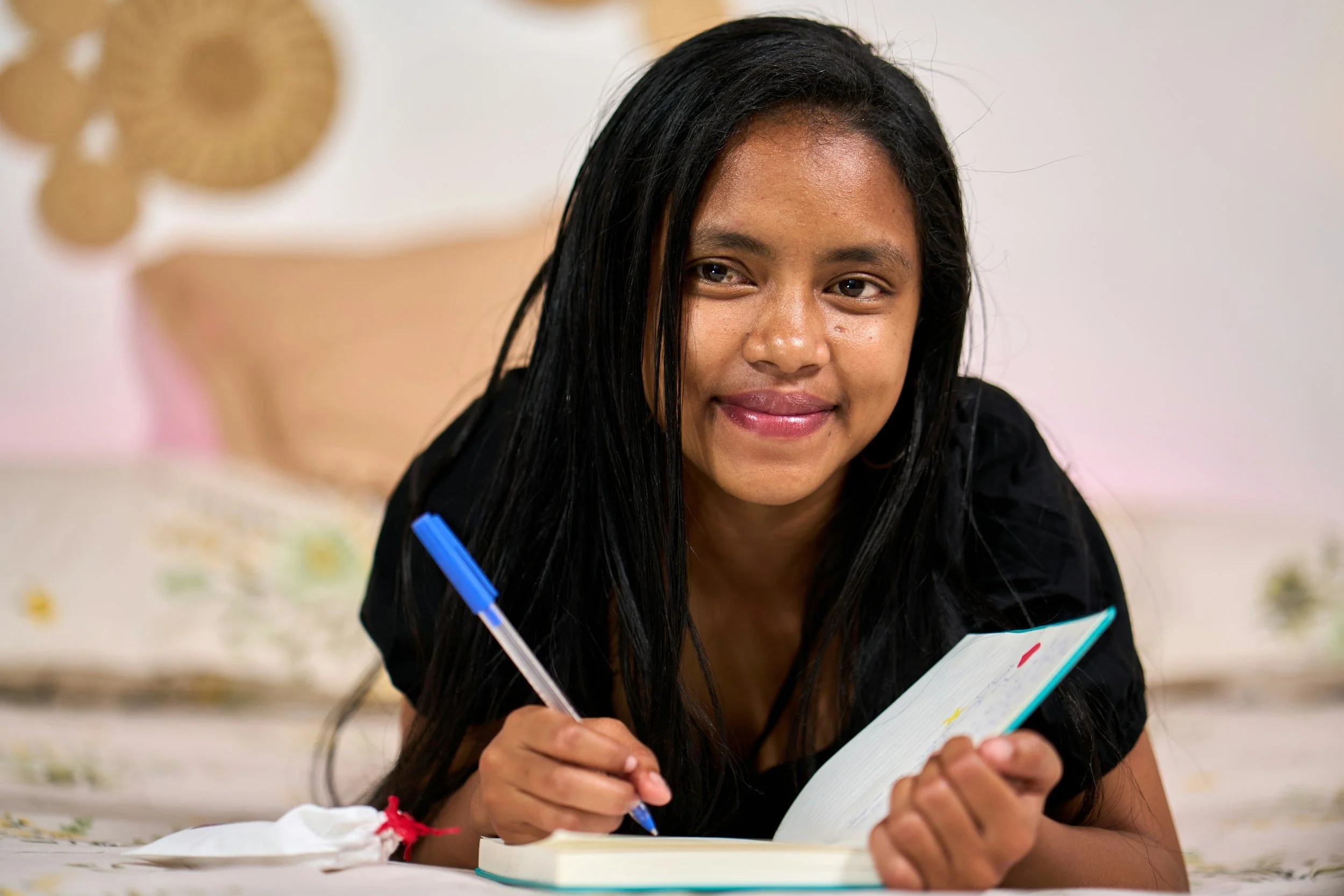 Young woman with long black hair smiling while writing in a notebook with a blue pen.