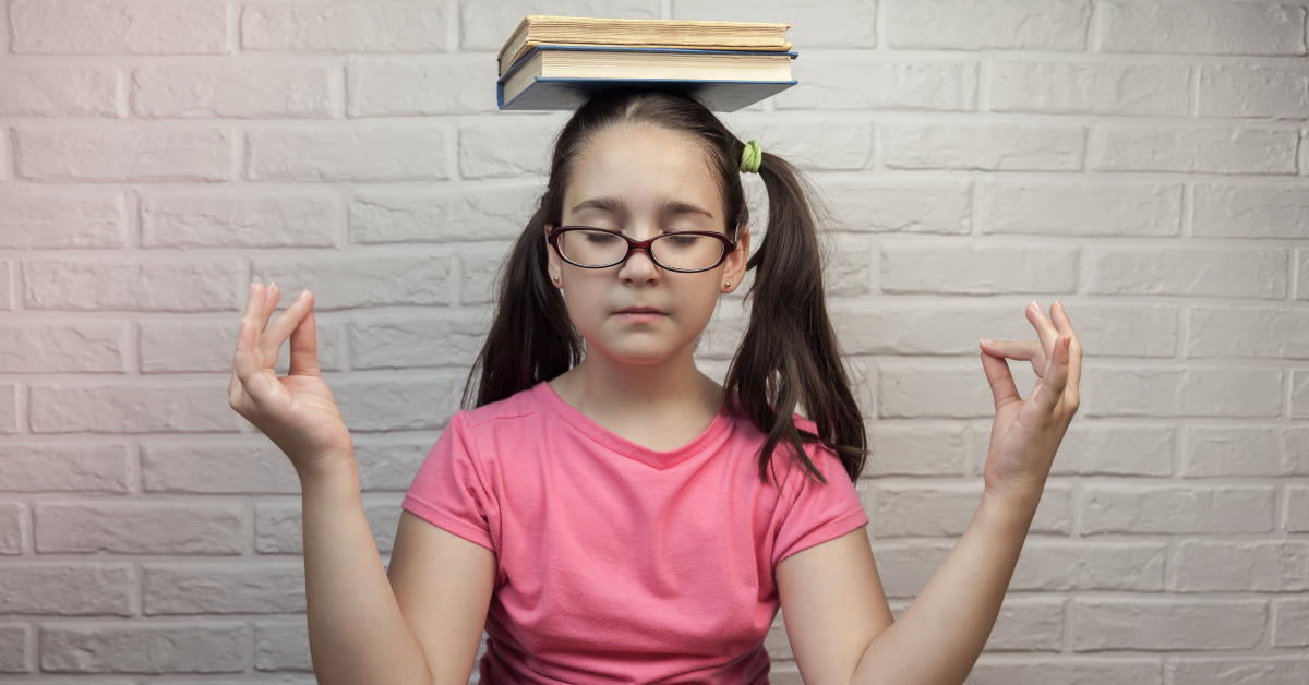 Girl with glasses and pigtails sitting with eyes closed, balancing a stack of books on her head, in front of a white brick wall.