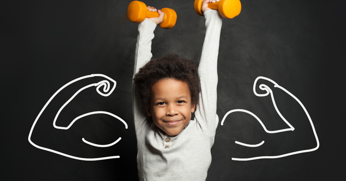 Young boy lifting orange dumbbells above his head in front of a blackboard with white drawing of strong arm muscles.