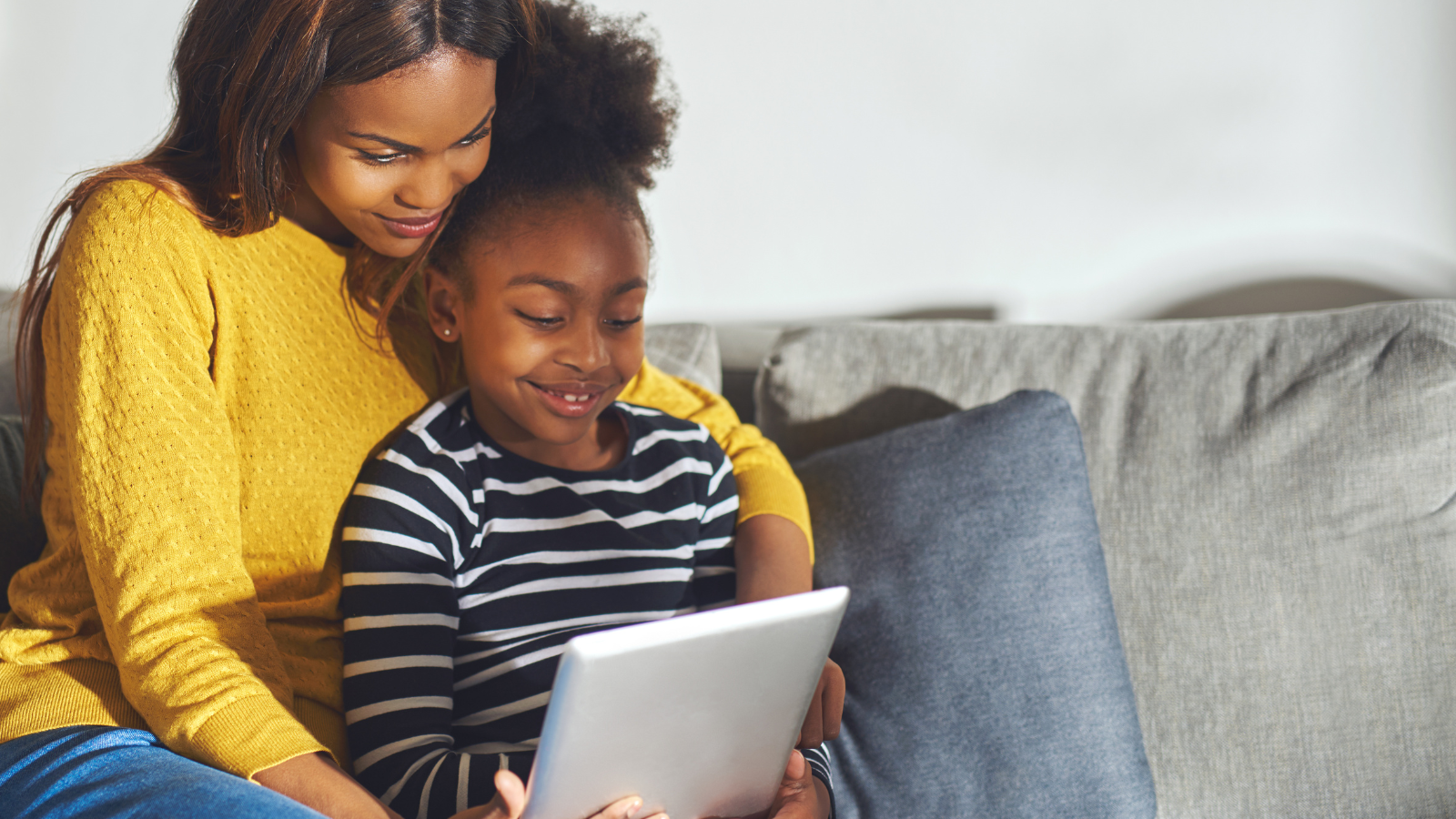 A woman and a girl sitting on a couch, looking at a tablet together, smiling.