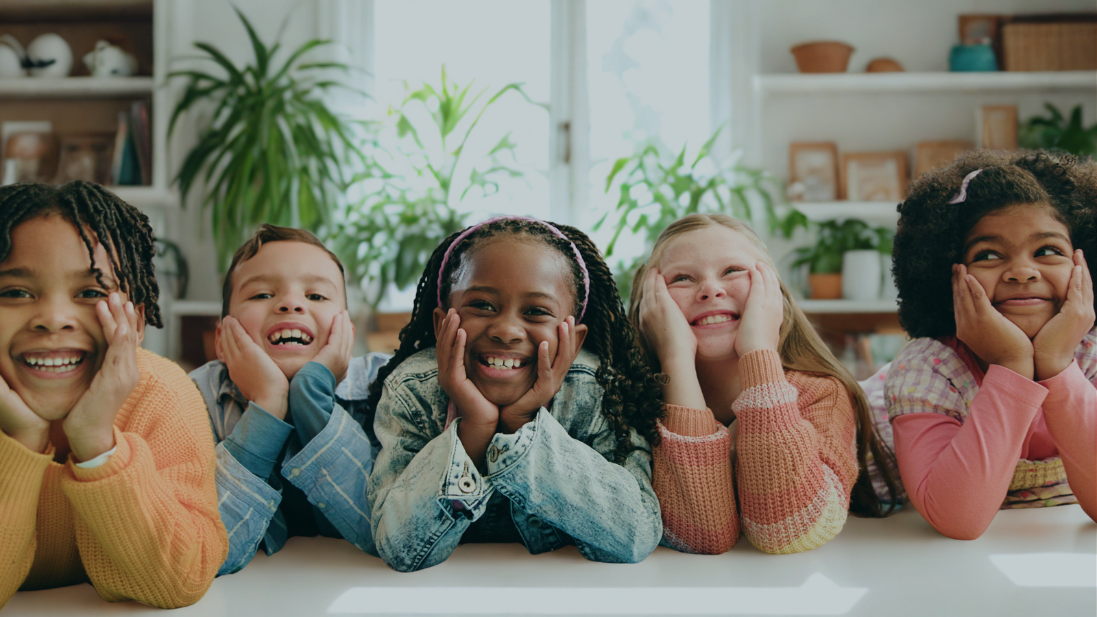 Five children smiling and resting their chins on their hands in a bright room.