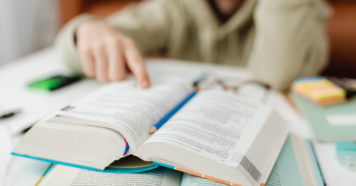 Open book with a blue pencil resting inside, on a cluttered desk with various stationery items.