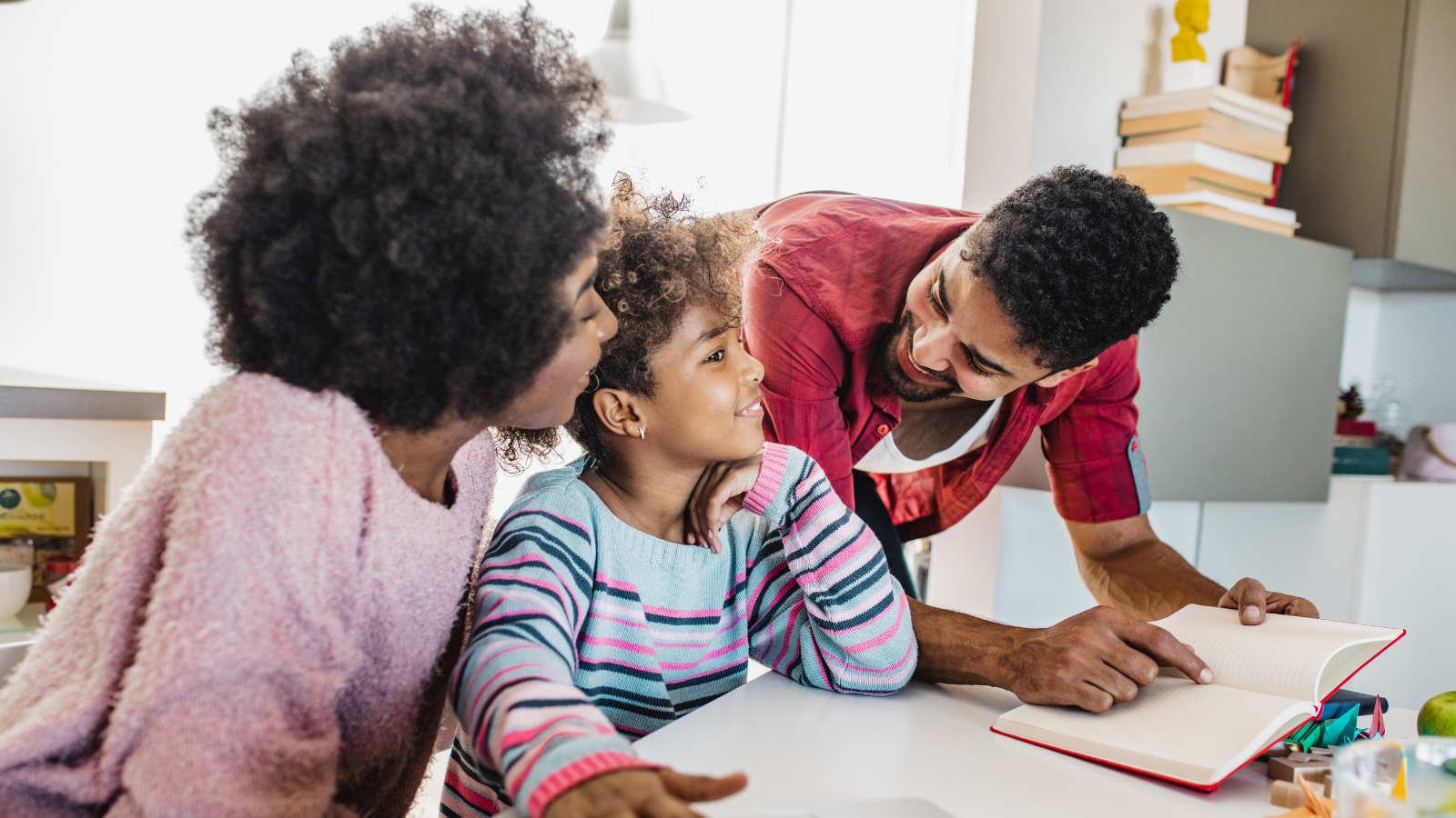 A family of three gathered around a table, smiling and engaging in a discussion, with a notebook open in front of them.