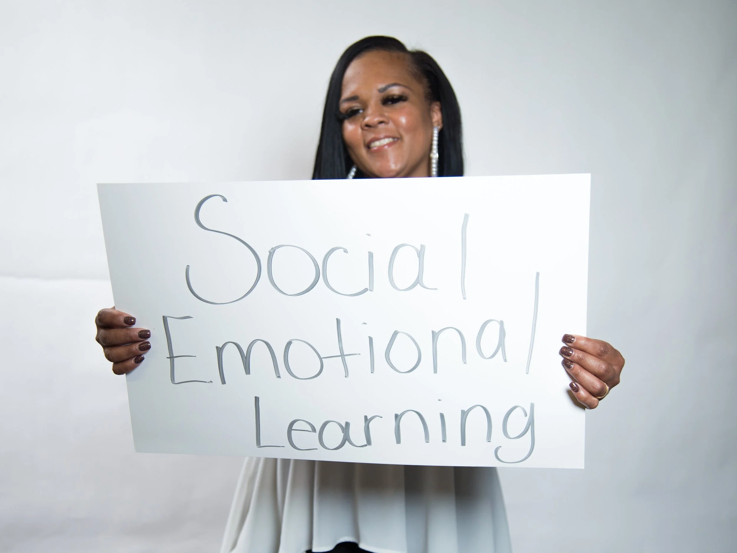 A woman with long black hair and earrings holding a sign that reads 'Social Education Learning' in black cursive letters in front of a plain white background.