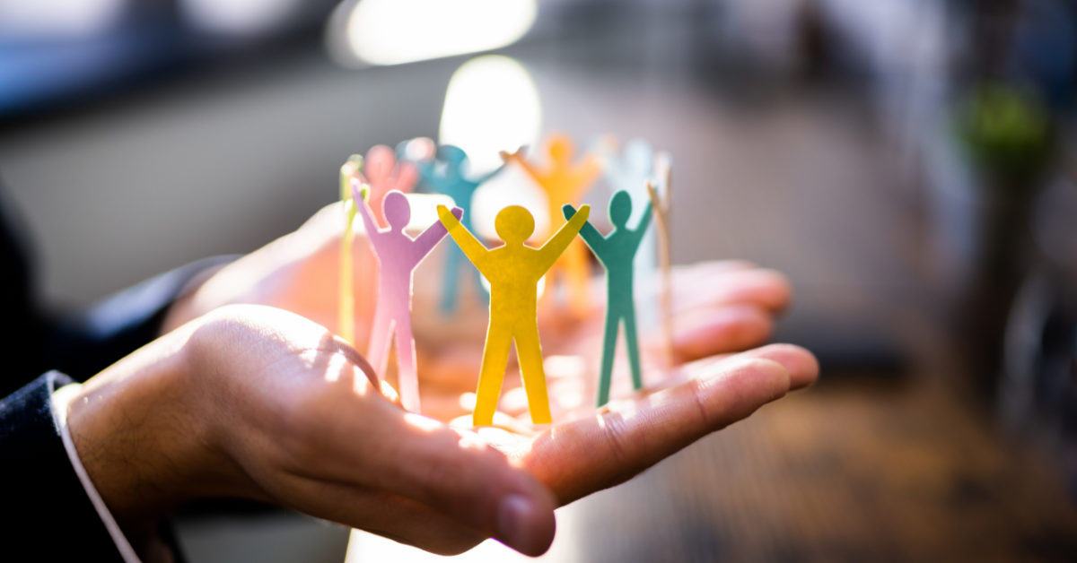 Hand holding colorful paper cutouts of people with arms raised, with a blurred background of a birthday cake with candles.