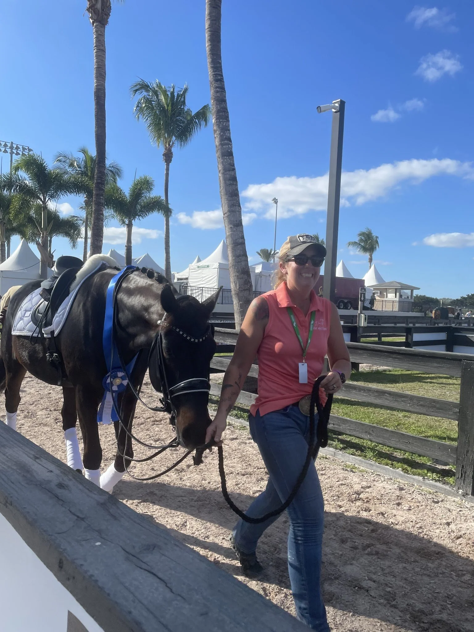 A woman in a pink sleeveless shirt, jeans, sunglasses, and a cap walking a horse on a dirt path, with palm trees, tents, and a blue sky in the background.