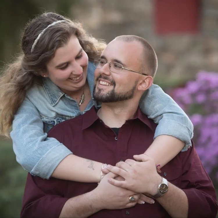 A smiling woman hugging a man from behind in an outdoor setting with purple flowers in the background.
